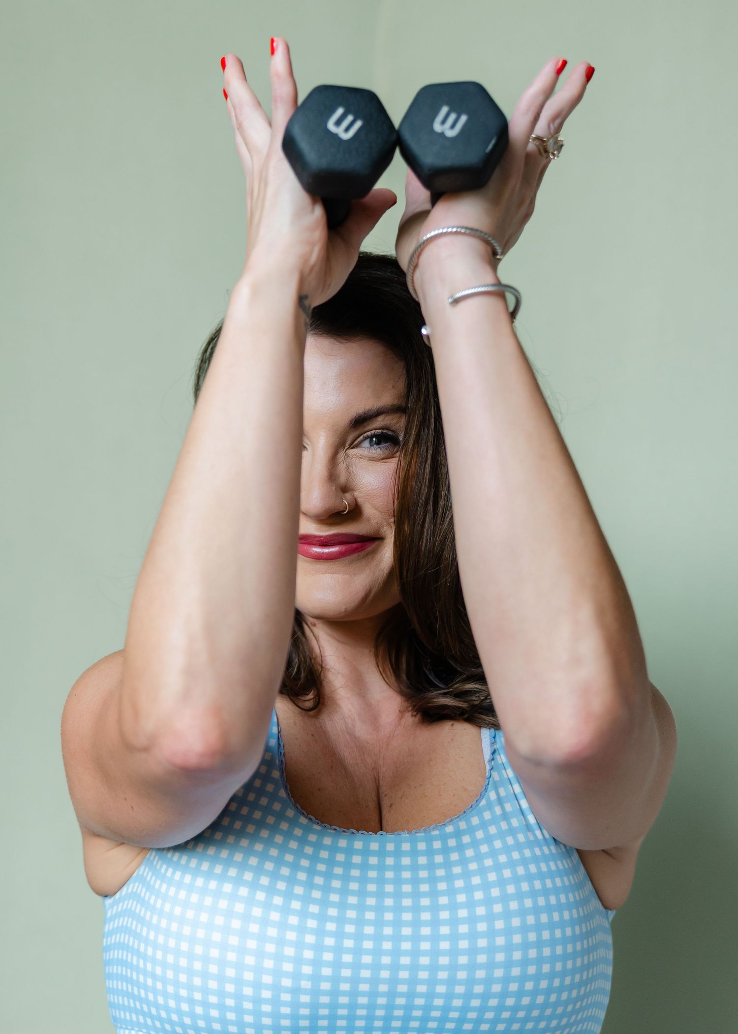 A woman in a blue patterned swimsuit lifts two dumbbells overhead against a green background.