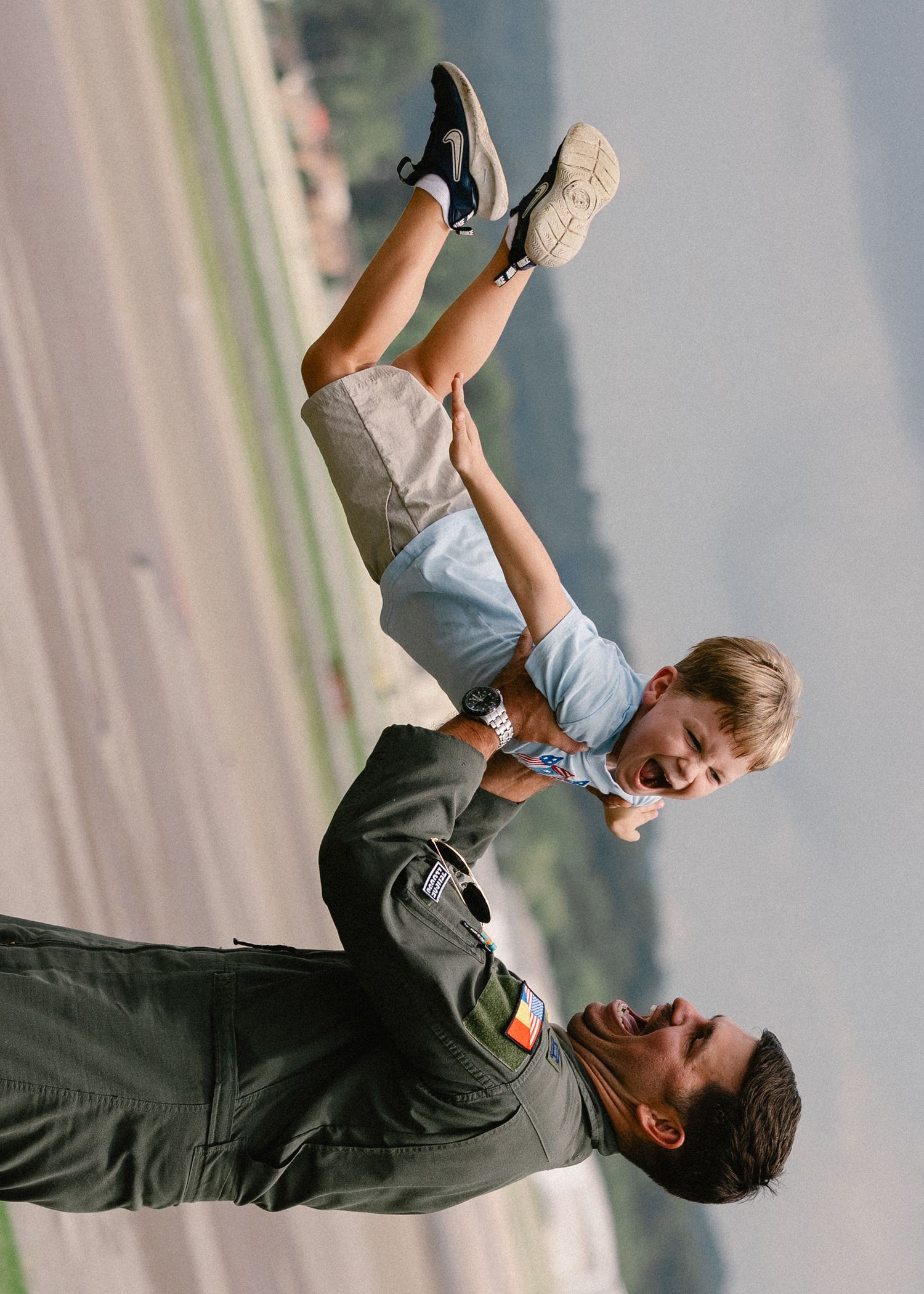 Smiling military pilot in green flight suit playfully holds laughing child upside down on an airfield runway.