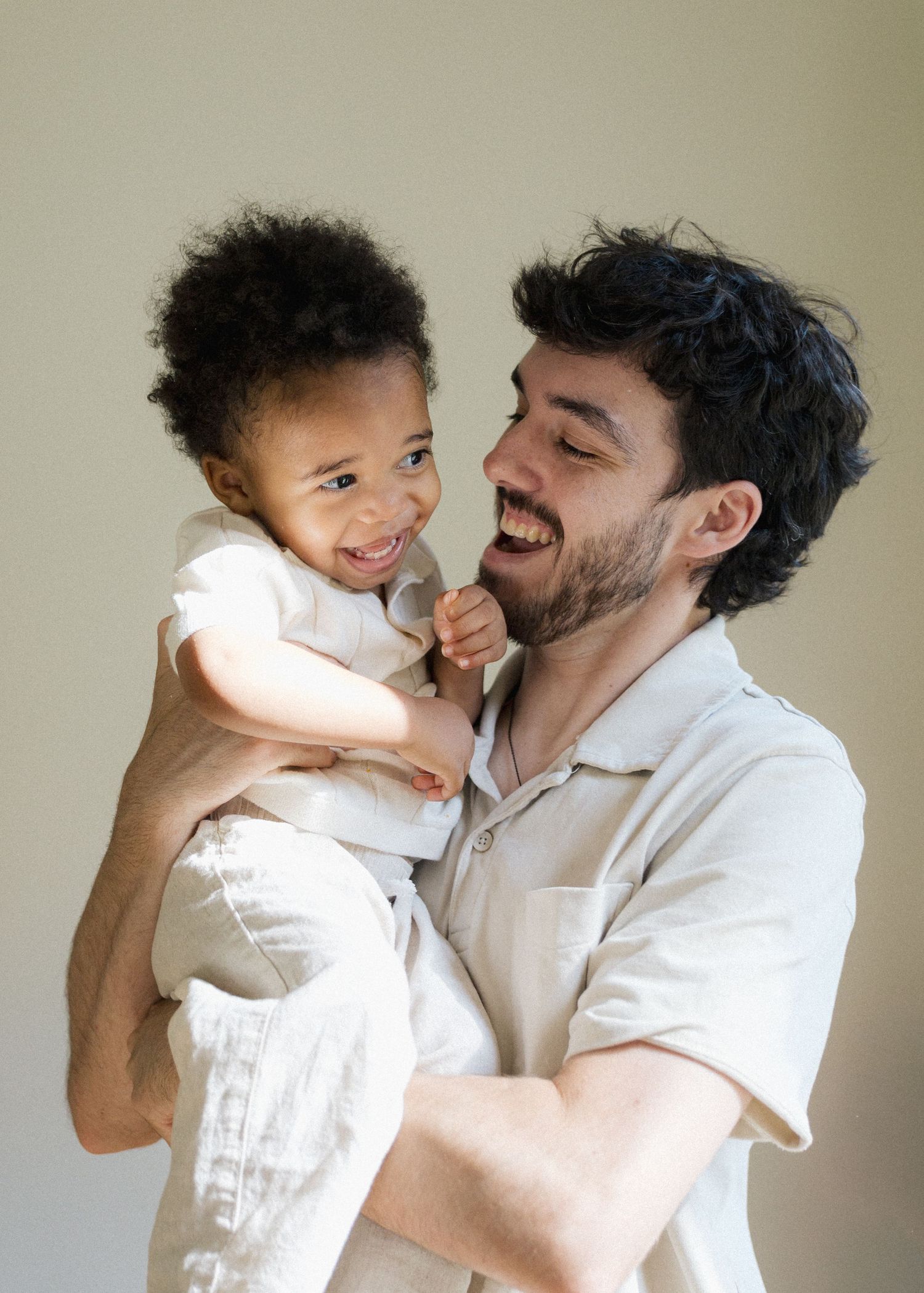 Laughing father holding his curly-haired toddler in a joyful indoor portrait.