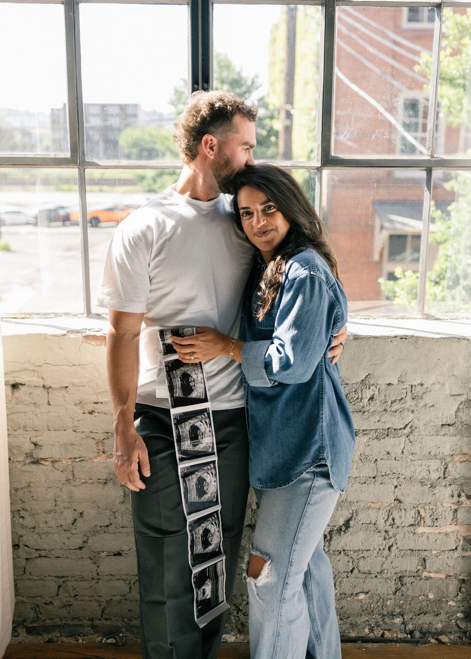 Couple embracing tenderly by a brick wall window holding ultrasound sonogram photos during pregnancy reveal.
