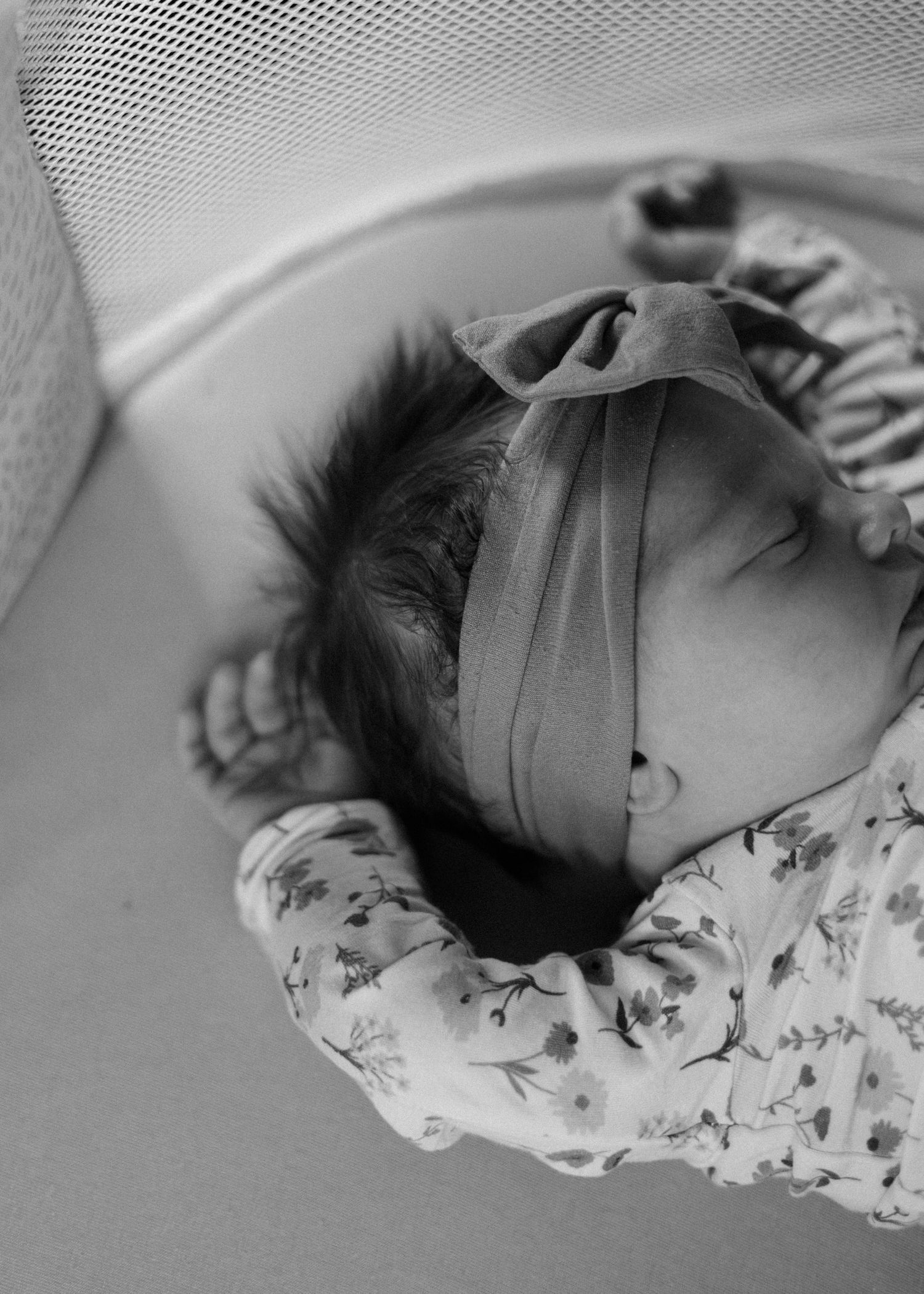 Sleeping newborn baby in a floral outfit and knotted headband lying in a mesh-sided bassinet in black and white.