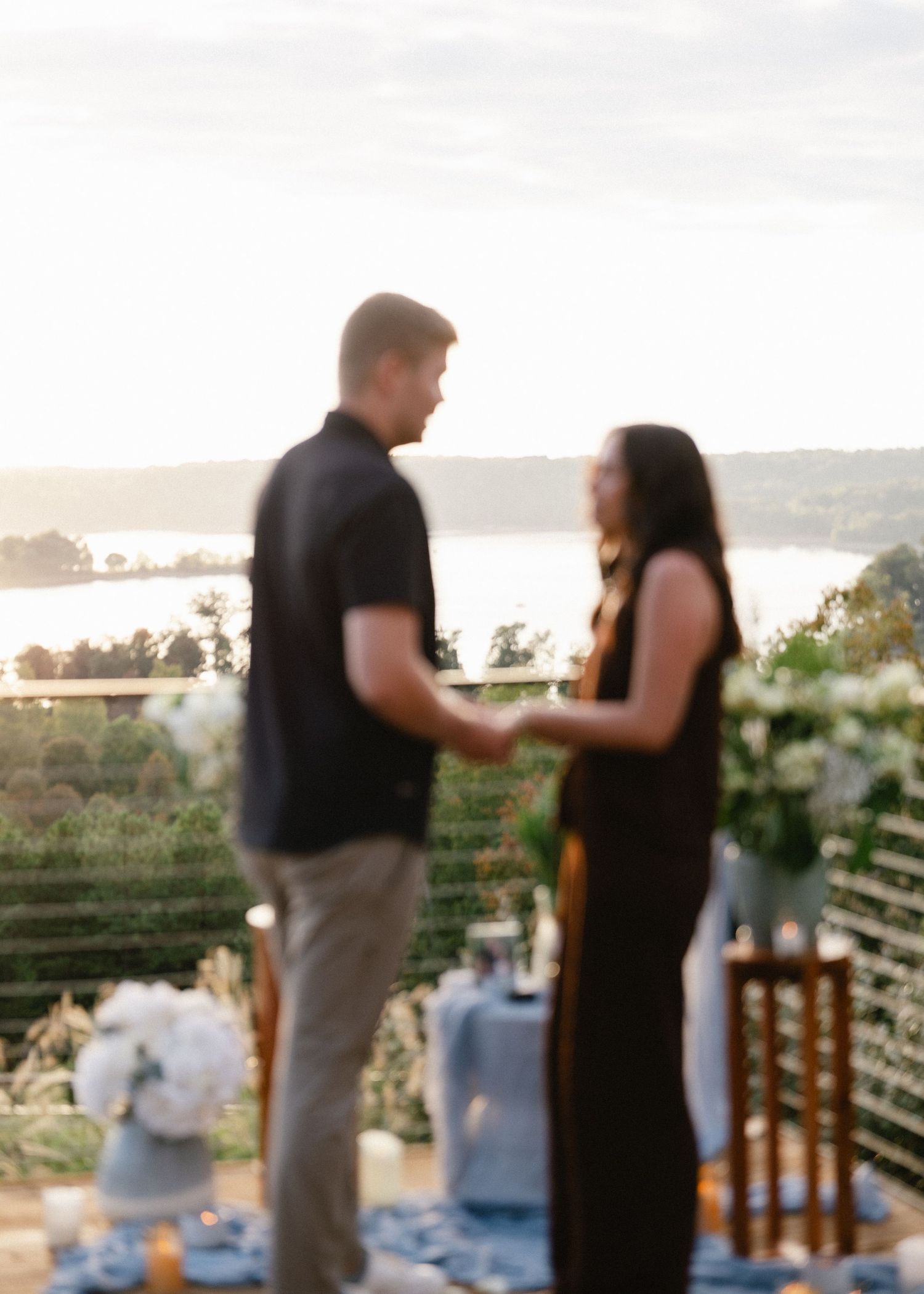 Couple holding hands at a romantic outdoor proposal setup with candles and white flowers overlooking a scenic lake at sunset.