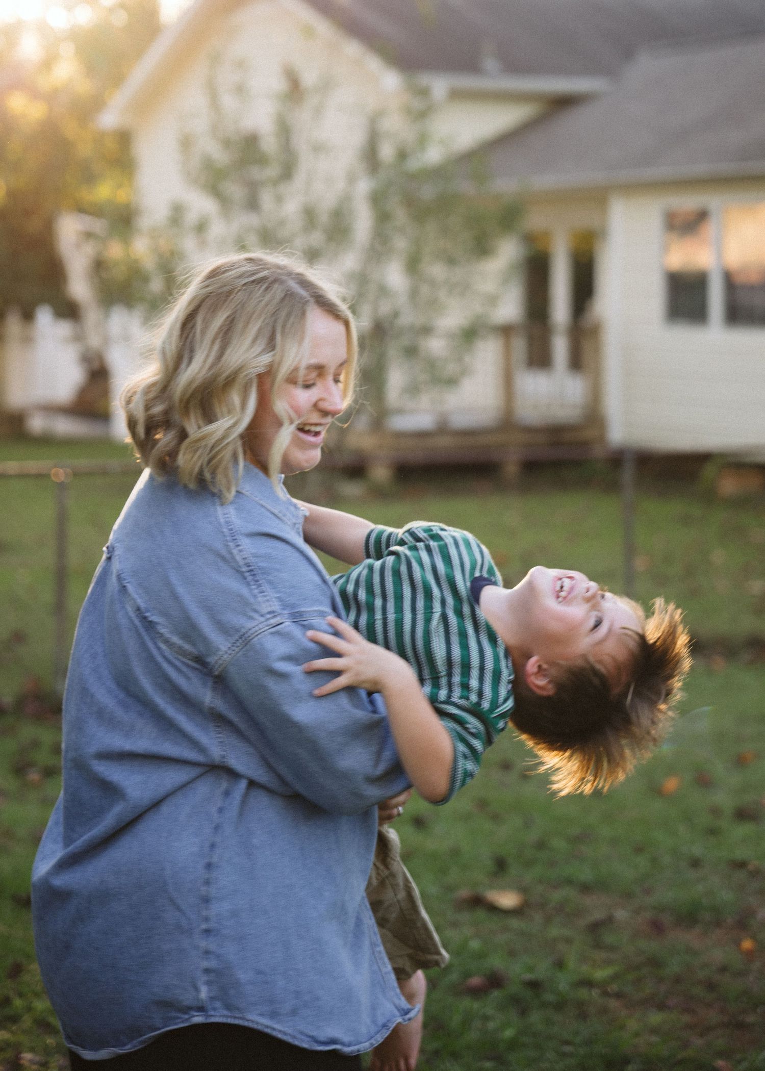 Smiling woman in a denim shirt swings a laughing child with tousled hair in a sunlit backyard with a house in the background.