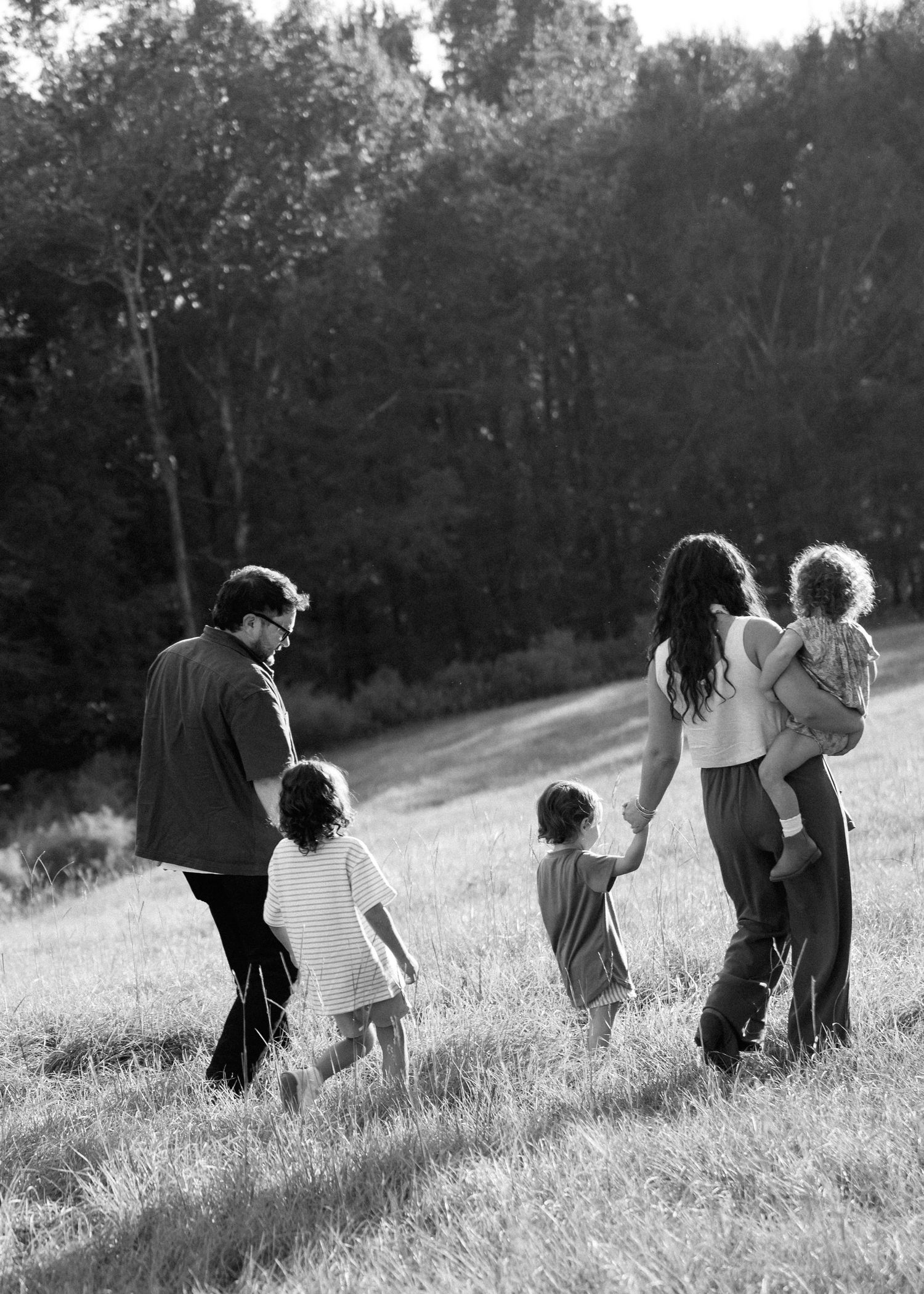 Black and white photo of a family of five walking together through a sunlit grassy field toward a dense tree line.