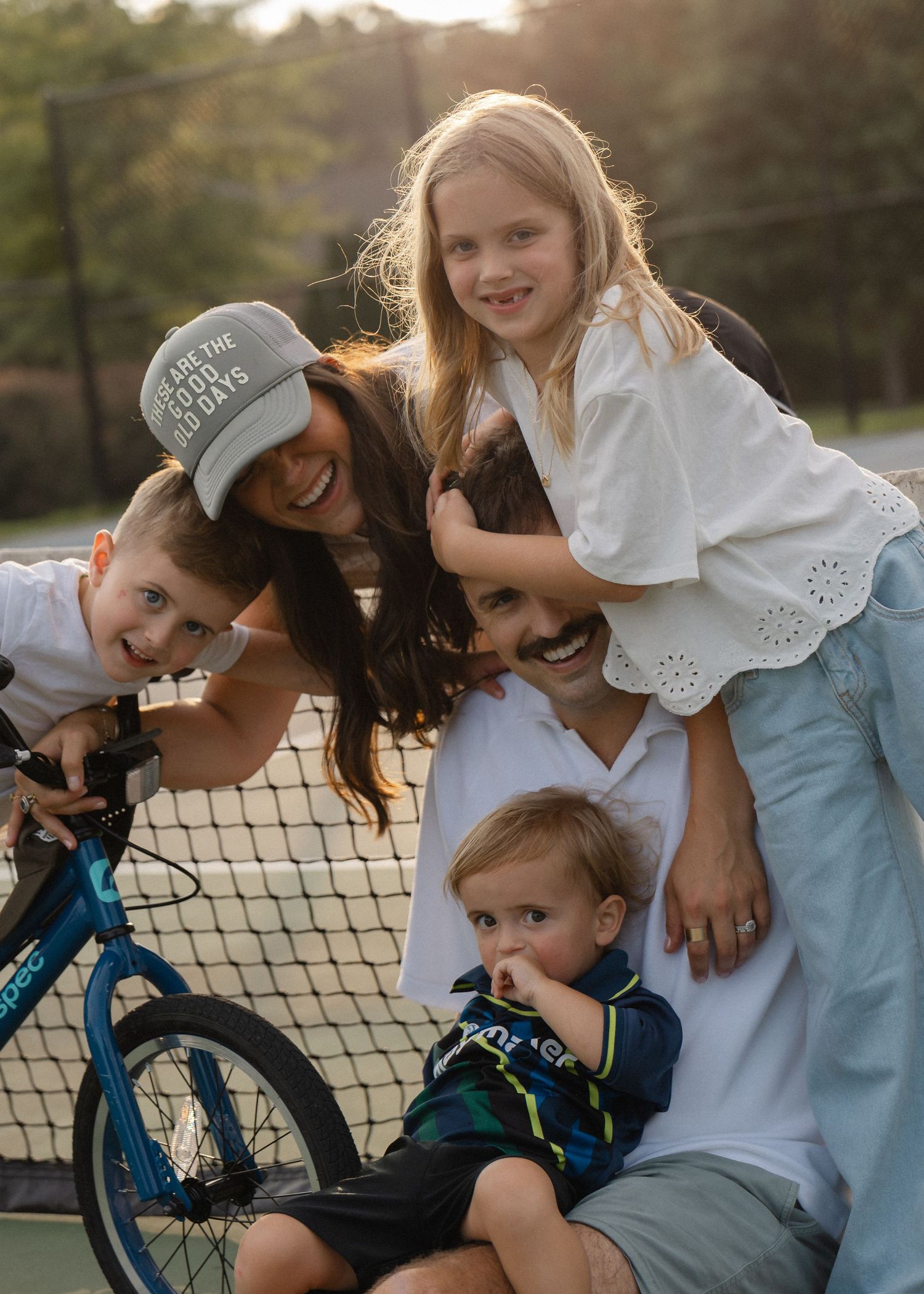 Happy family of five posing together on a tennis court at sunset, with a blue bicycle nearby.