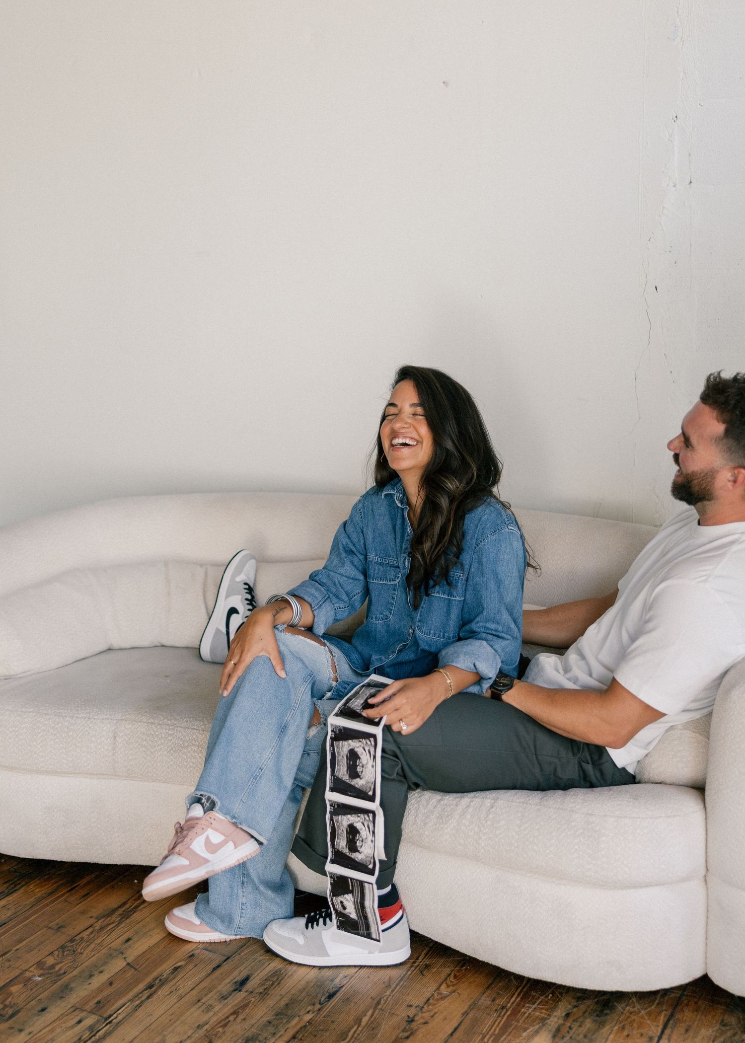 Joyful couple laughing together on a cream couch holding ultrasound sonogram photos in a bright studio.