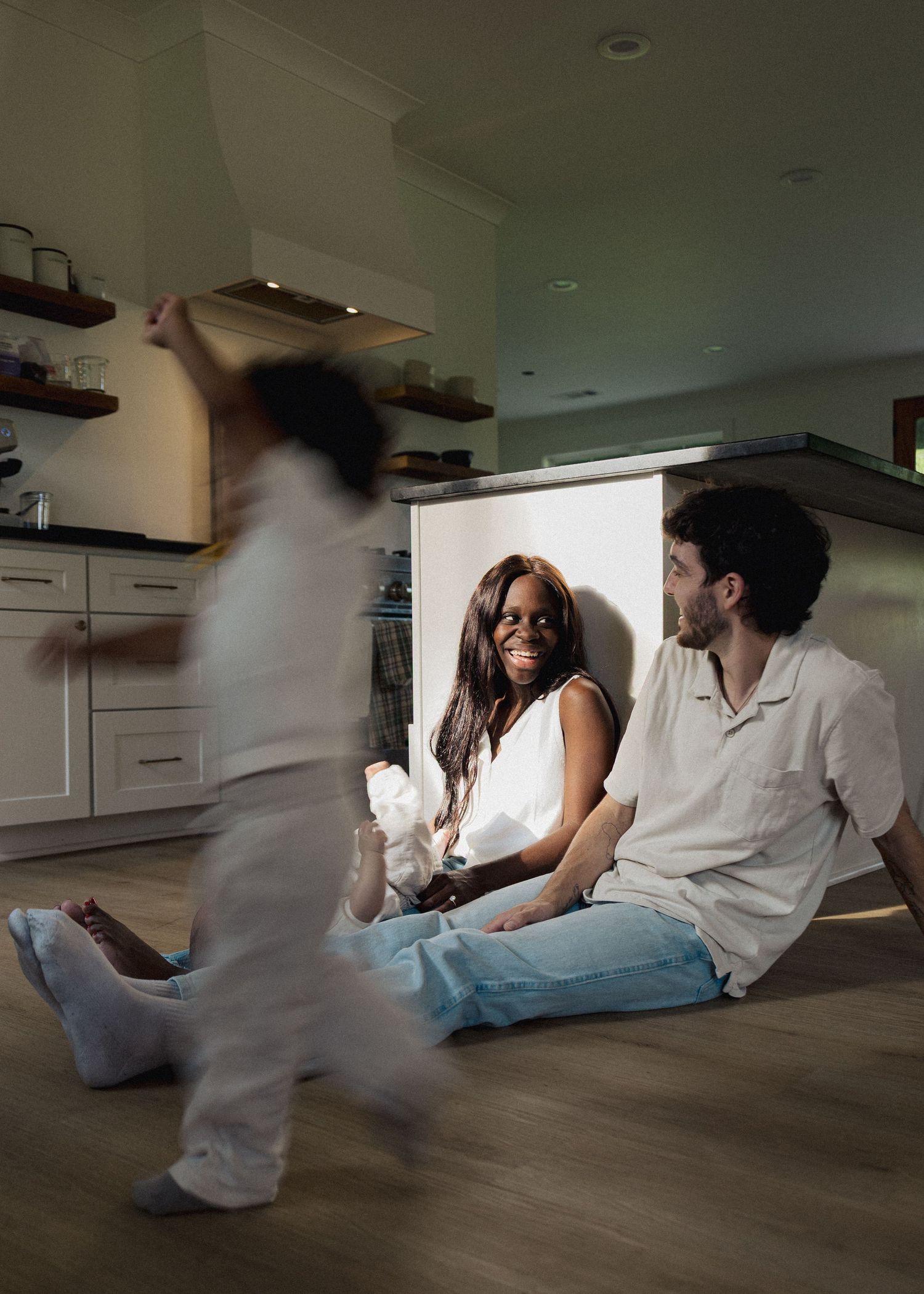 A happy interracial family sits on kitchen floor laughing while their child plays nearby.