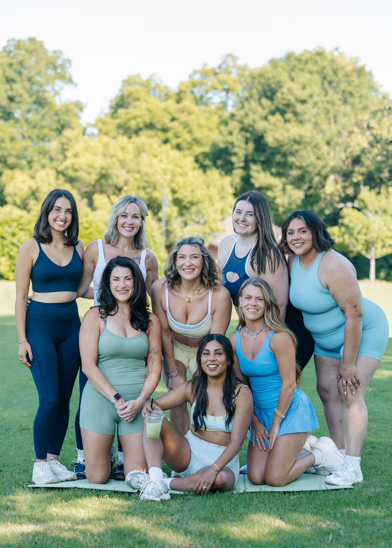 Group of nine smiling women in athletic wear posing together on green grass in a park.