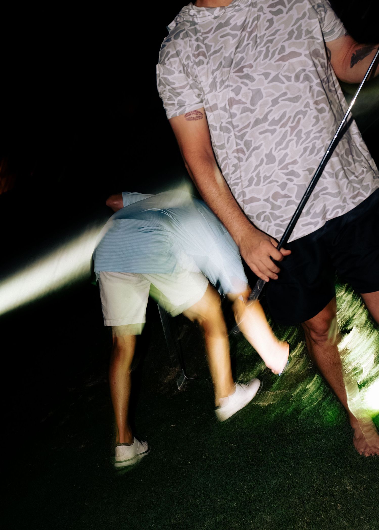 Two golfers walk along a dark golf course at night carrying clubs under a beam of light.