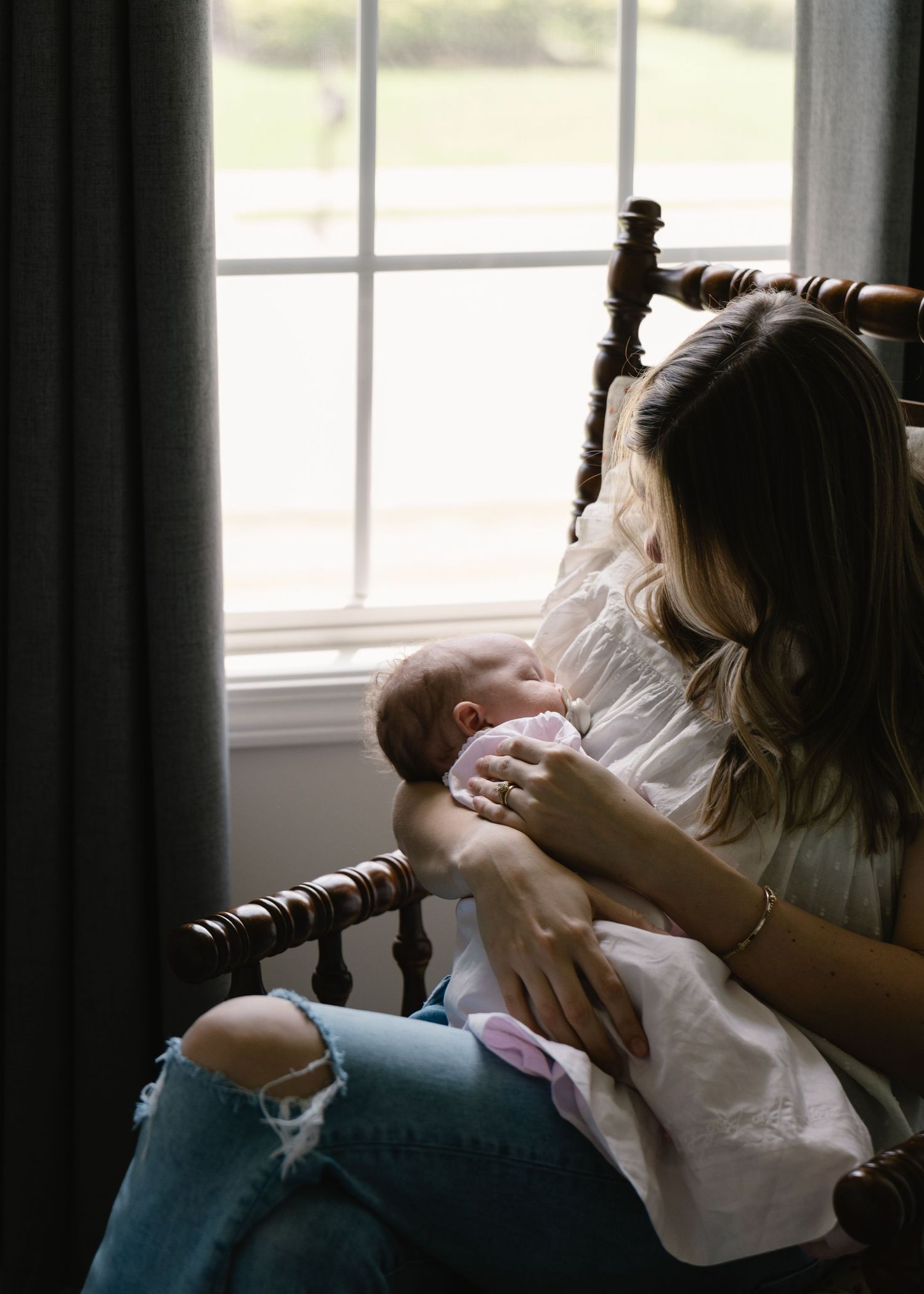 Mother sitting in wooden rocking chair by window cradling her newborn baby.