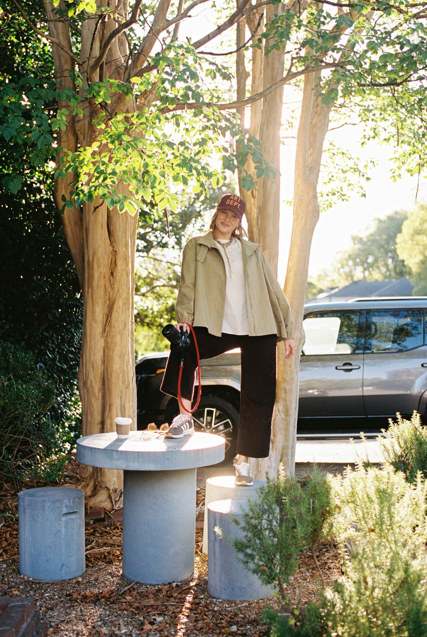 Person in a cap, olive jacket and black pants standing on a concrete stool beside a tree, with a gray SUV and shrubs behind.