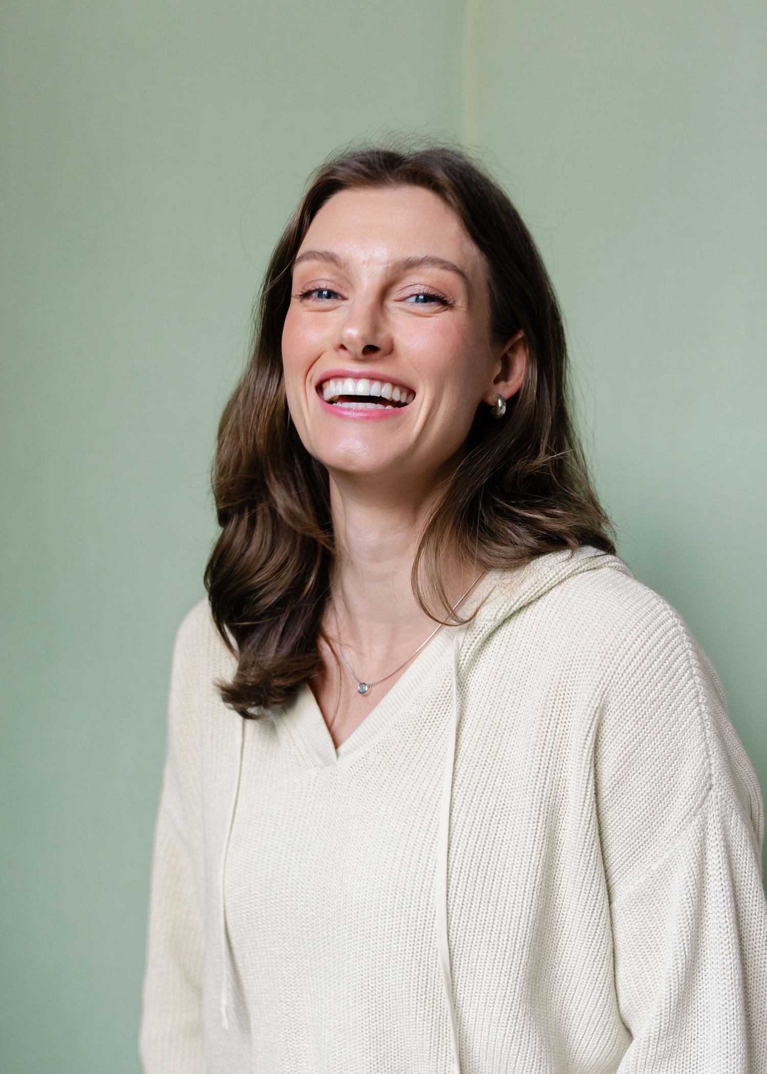 Brunette woman laughing in cream ribbed hoodie against sage green background.