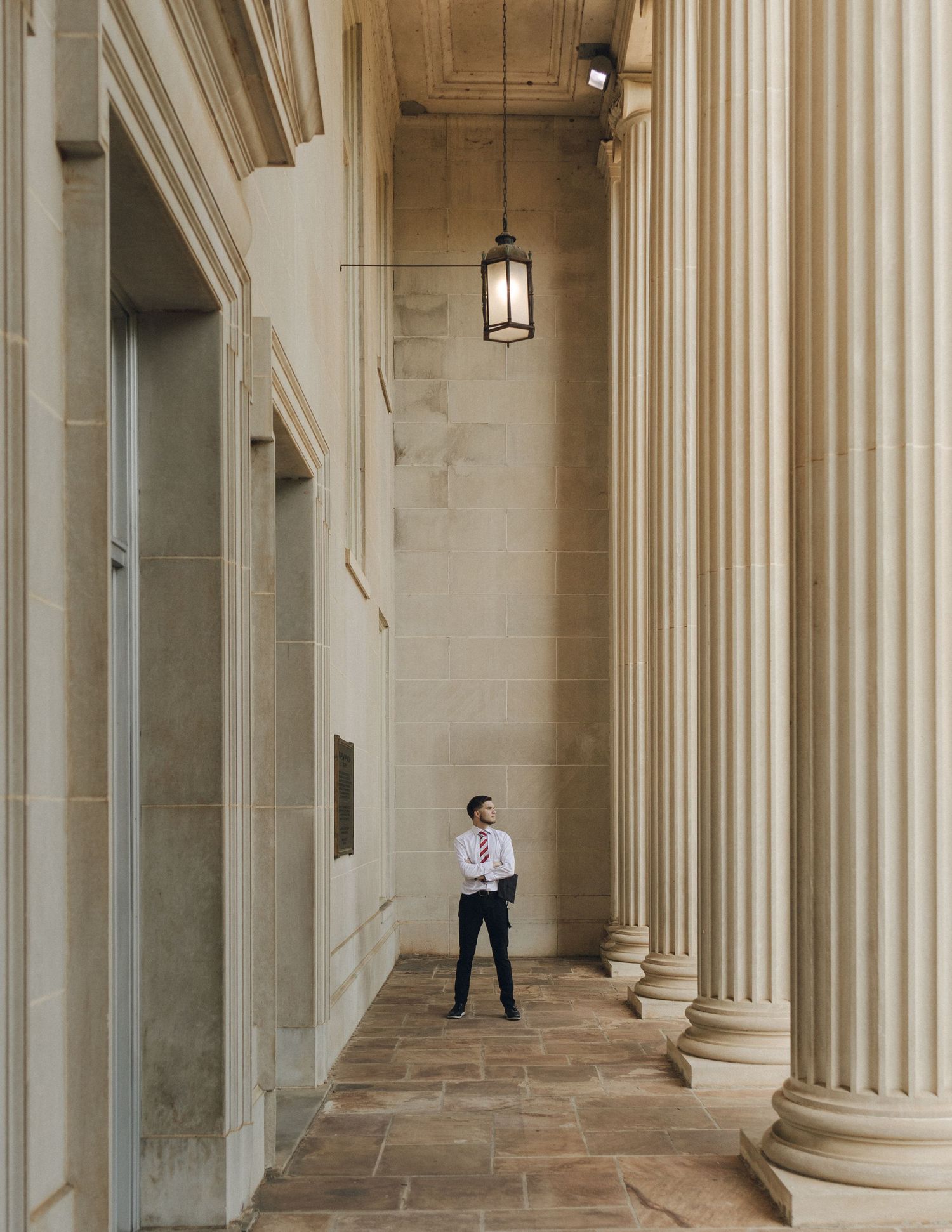 Man in dress shirt and tie standing among tall classical columns of grand stone building.