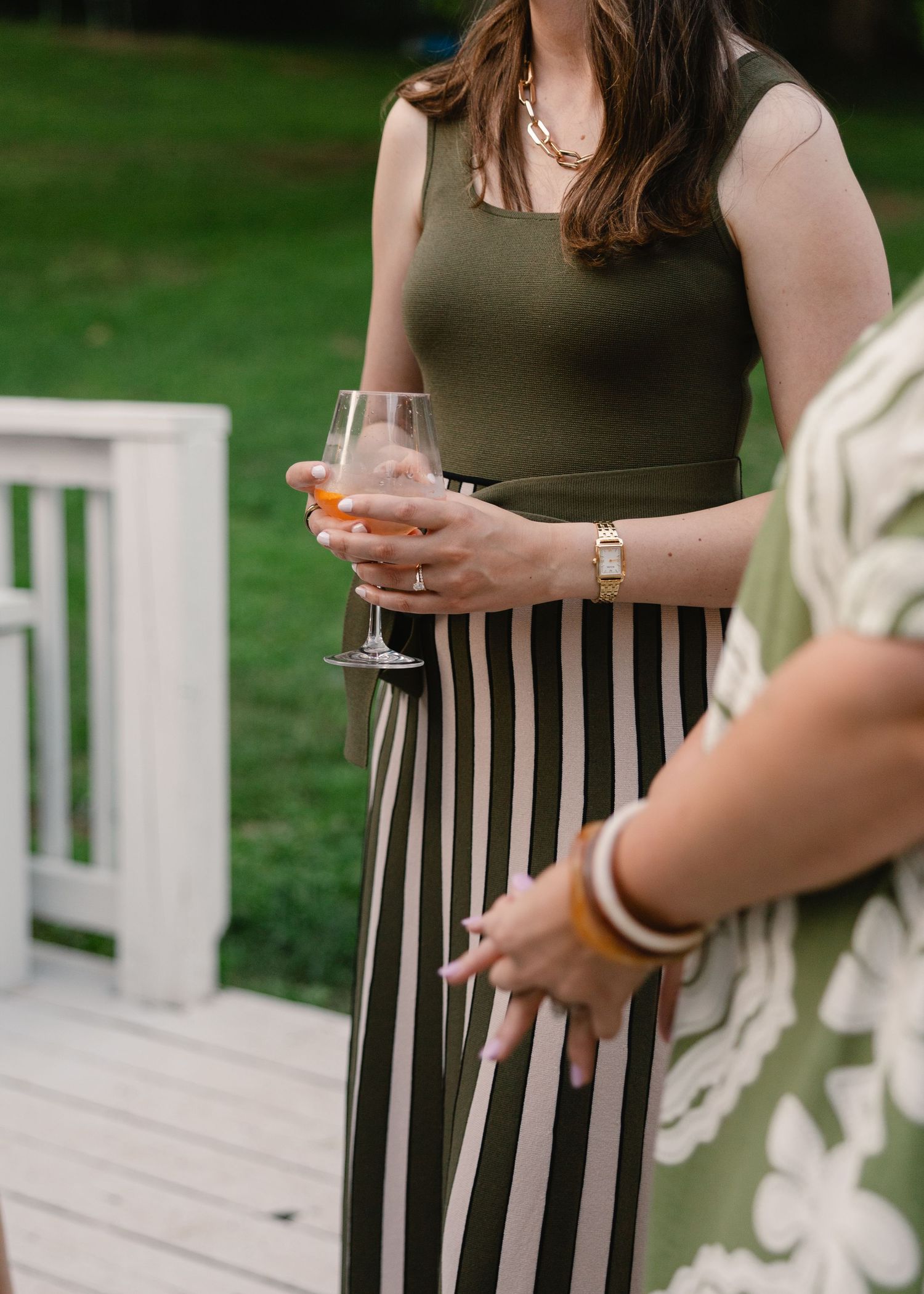 Woman in olive green top and striped skirt holding a rose wine glass at an outdoor summer gathering.