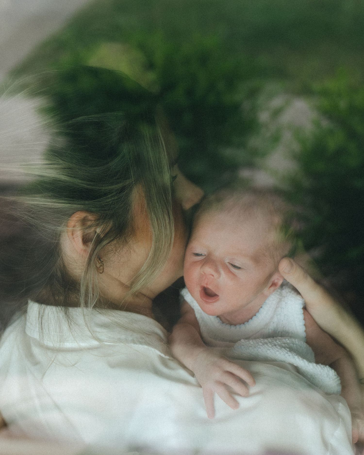 Mother gently kissing crying newborn baby captured through window glass.