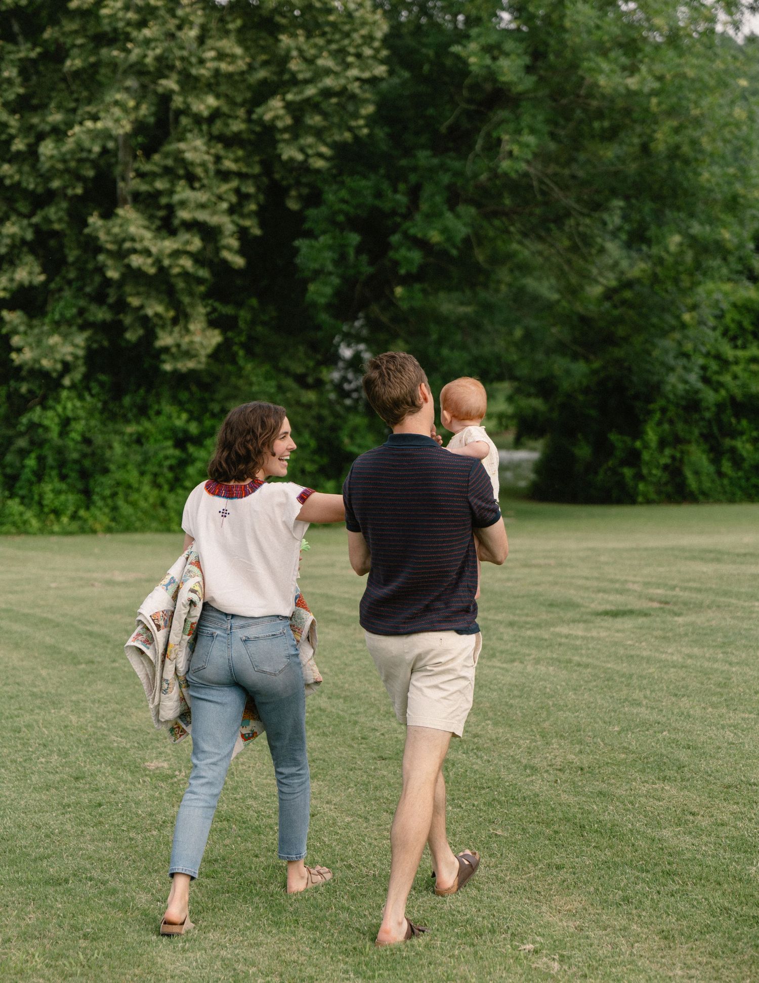 Young family walking across green park lawn with father carrying baby from behind.