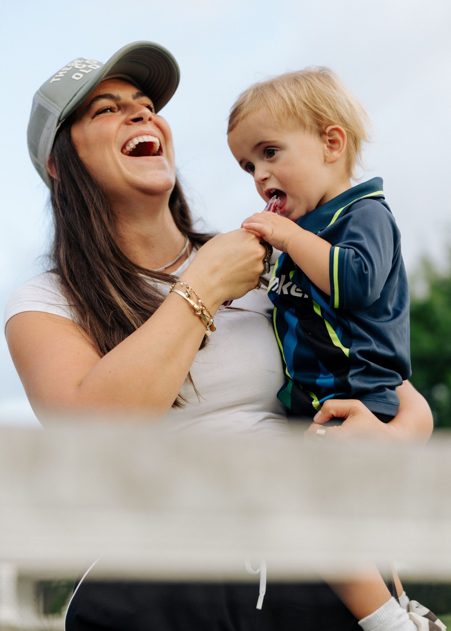 A laughing woman in a cap holds a toddler in a navy soccer jersey outdoors.