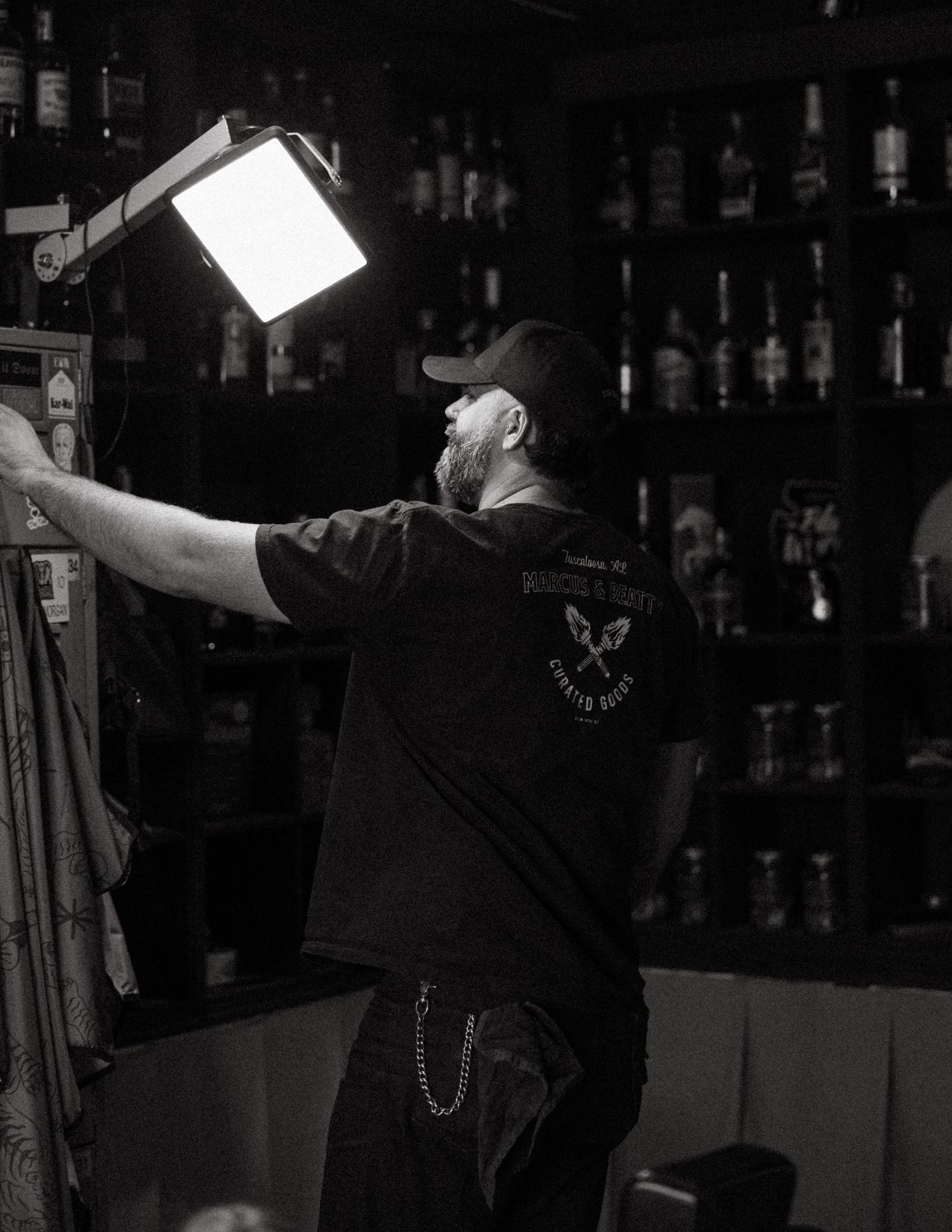 Black and white photo of a bearded man in a cap adjusting a light in a bar.