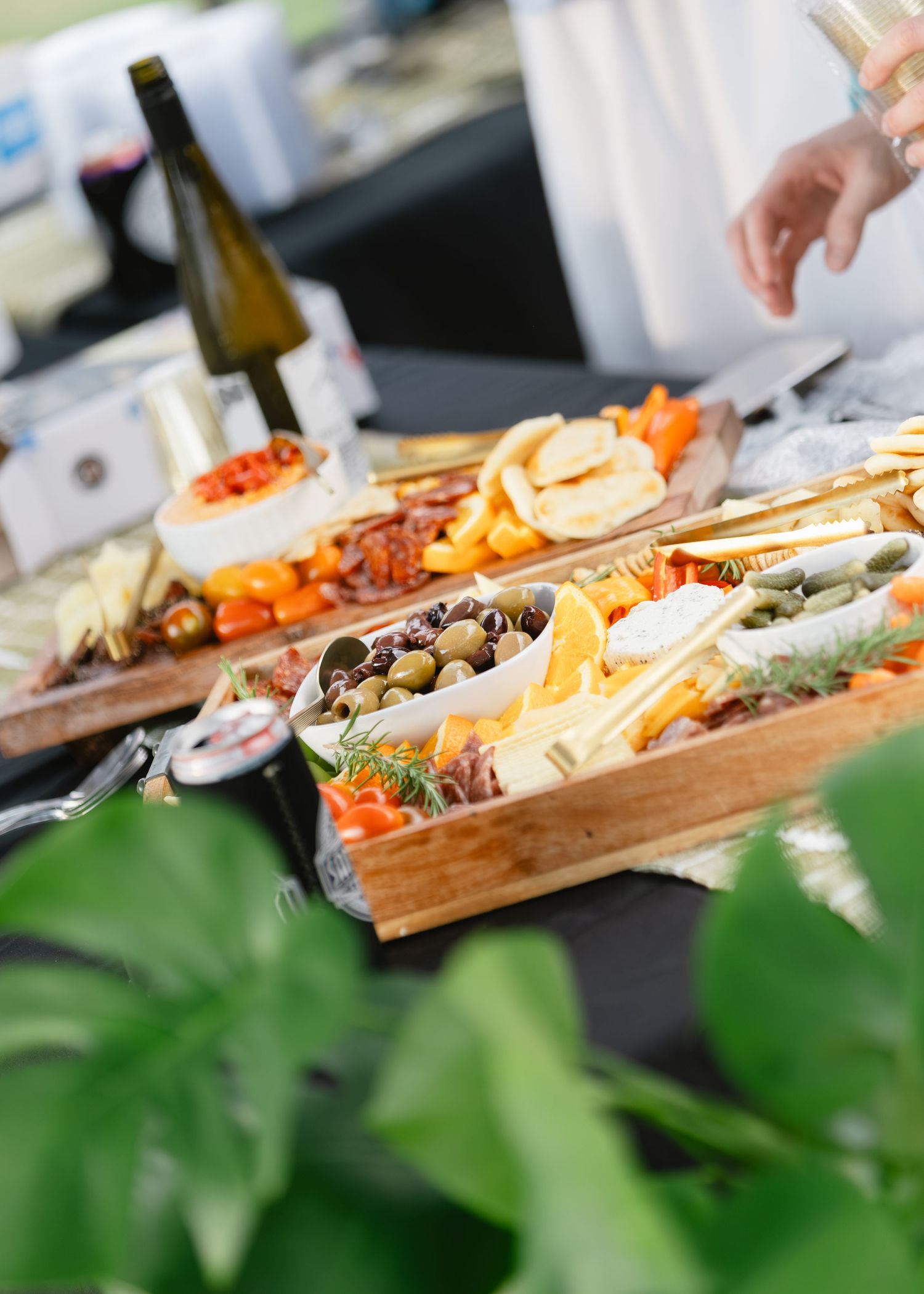 Wooden boards of catered charcuterie with cheeses, olives, cured meats and fruit beside a wine bottle as a guest reaches in.