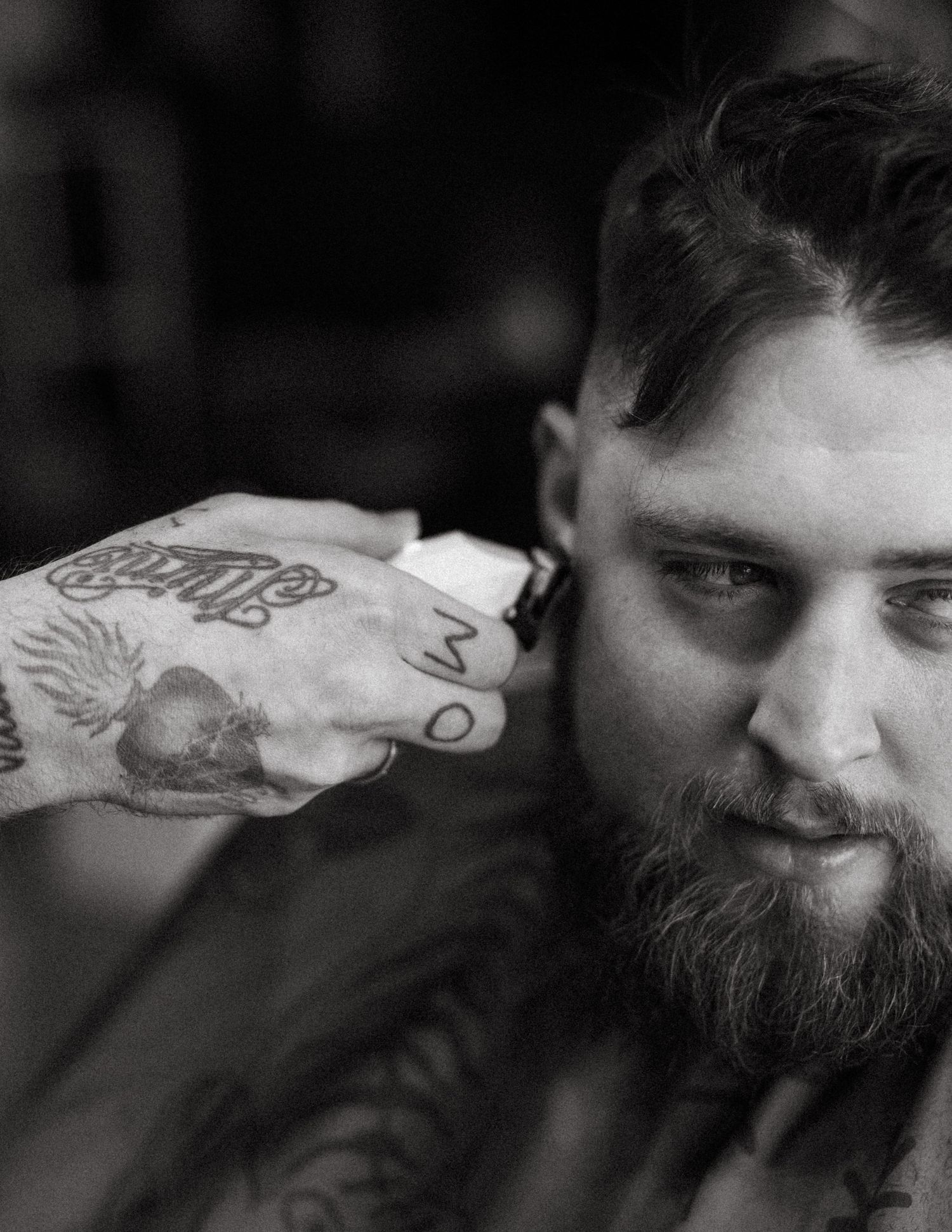 Black and white close-up of a heavily tattooed barber trimming a bearded client's hair.