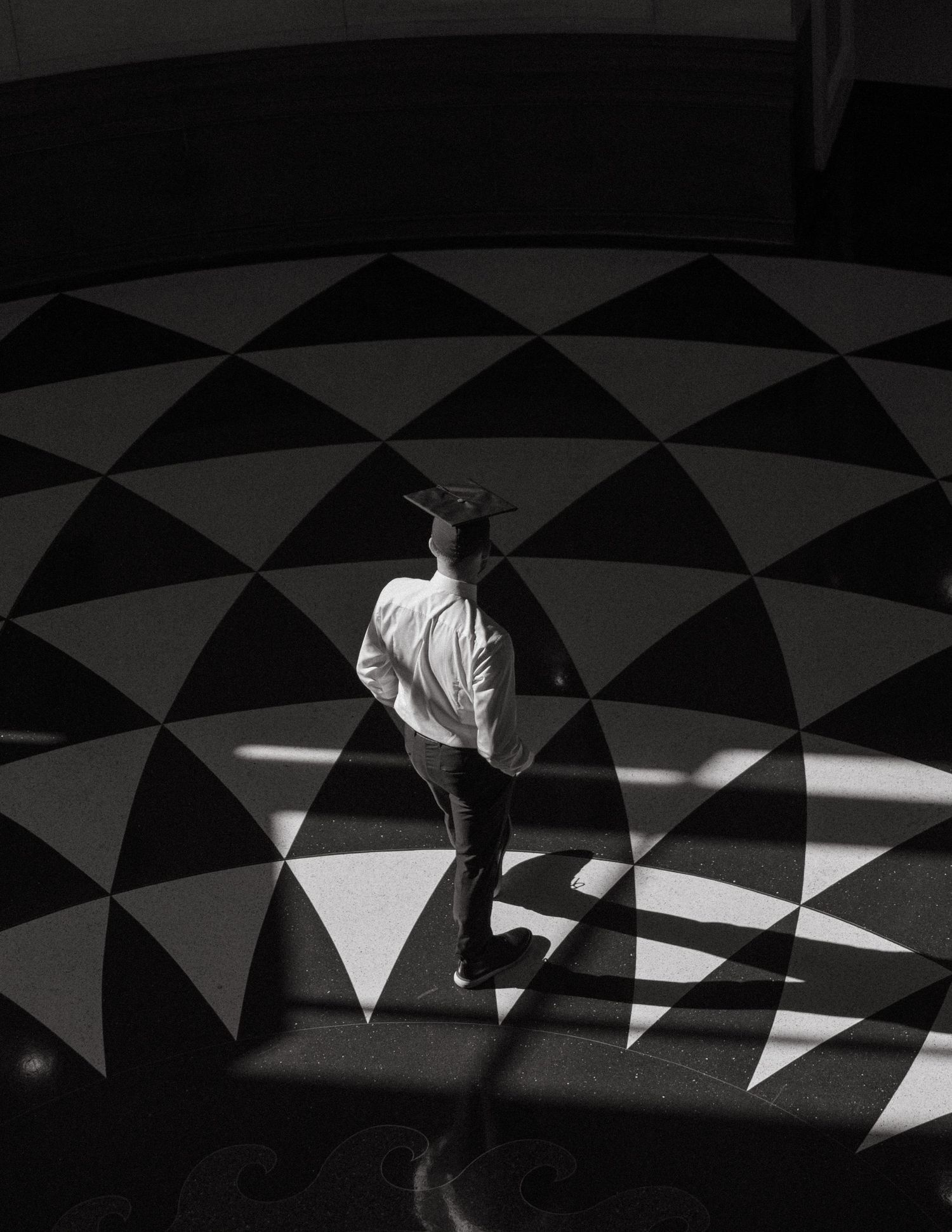Aerial black and white photo of person standing on geometric patterned floor with dramatic shadows.