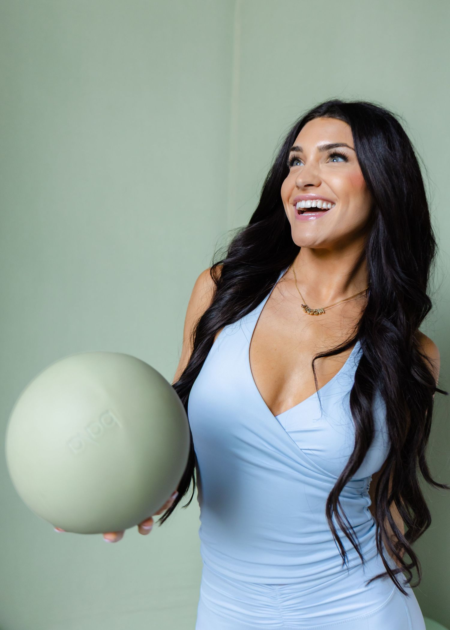 Smiling brunette woman in blue wrap top holding sage green pilates ball indoors.