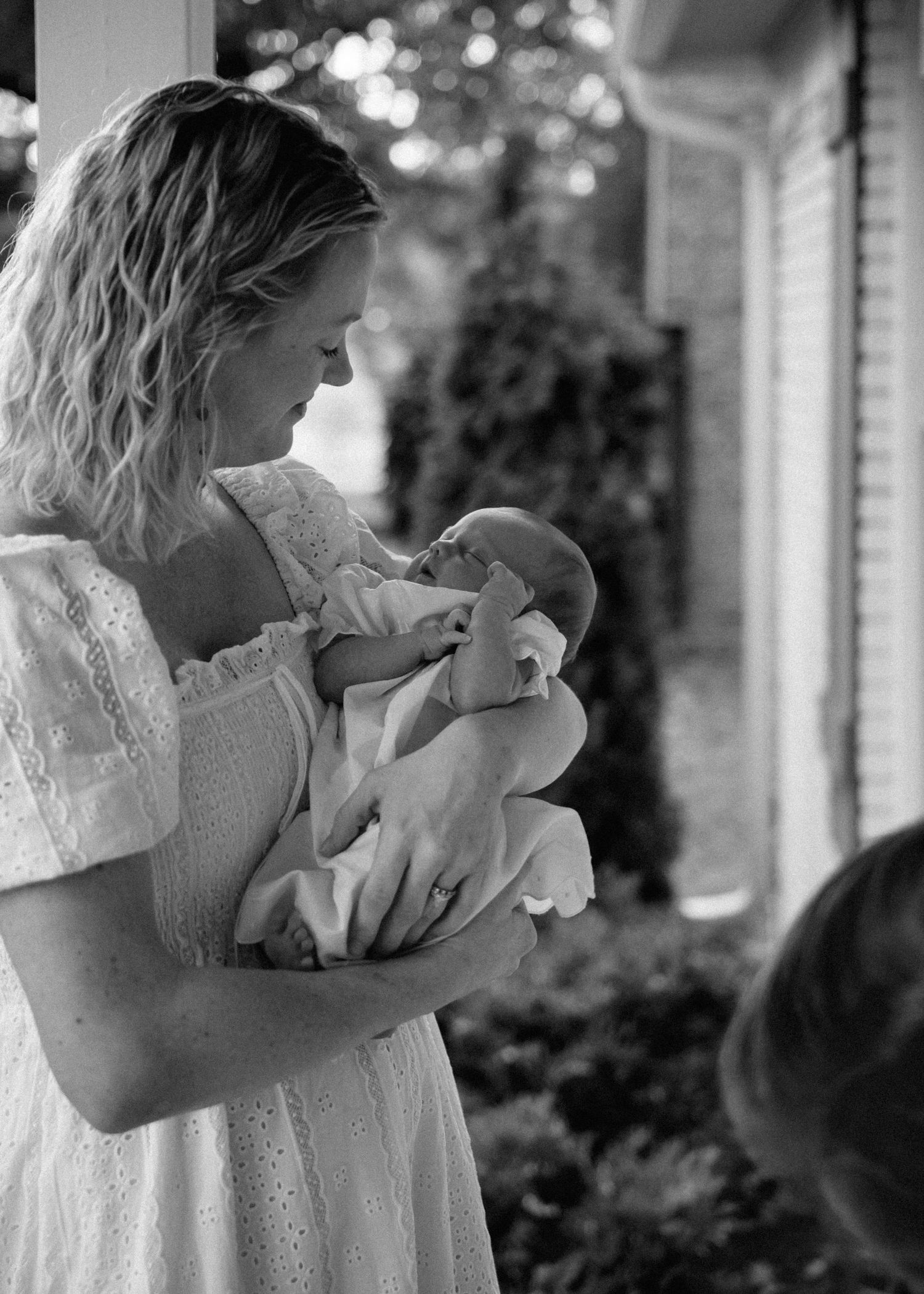 Black and white photo of a smiling mother in an eyelet dress gently holding her sleeping newborn baby outdoors near a house.