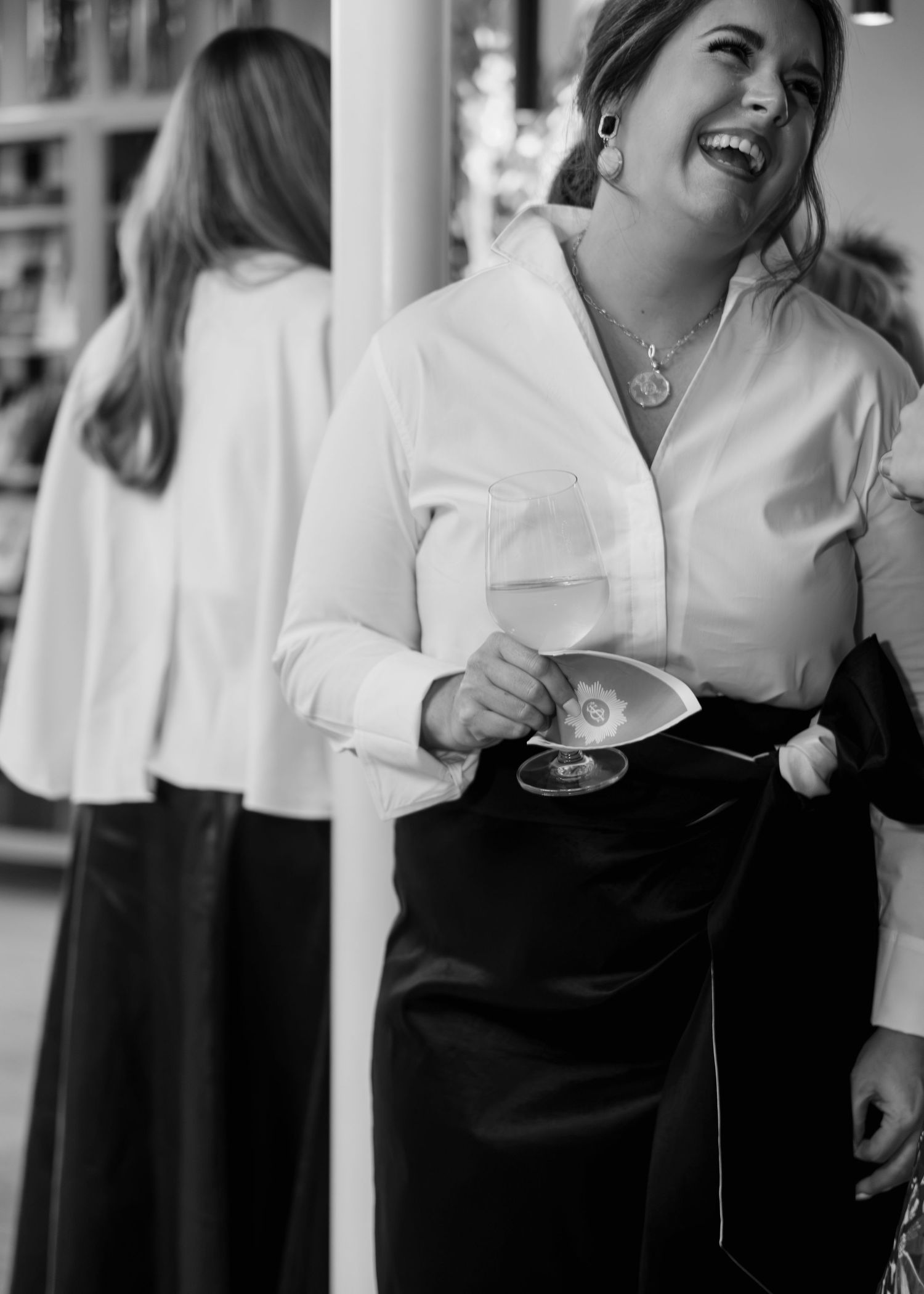 Black and white photo of a laughing woman in a white blouse and dark skirt holding a wine glass beside a mirror reflecting her back.