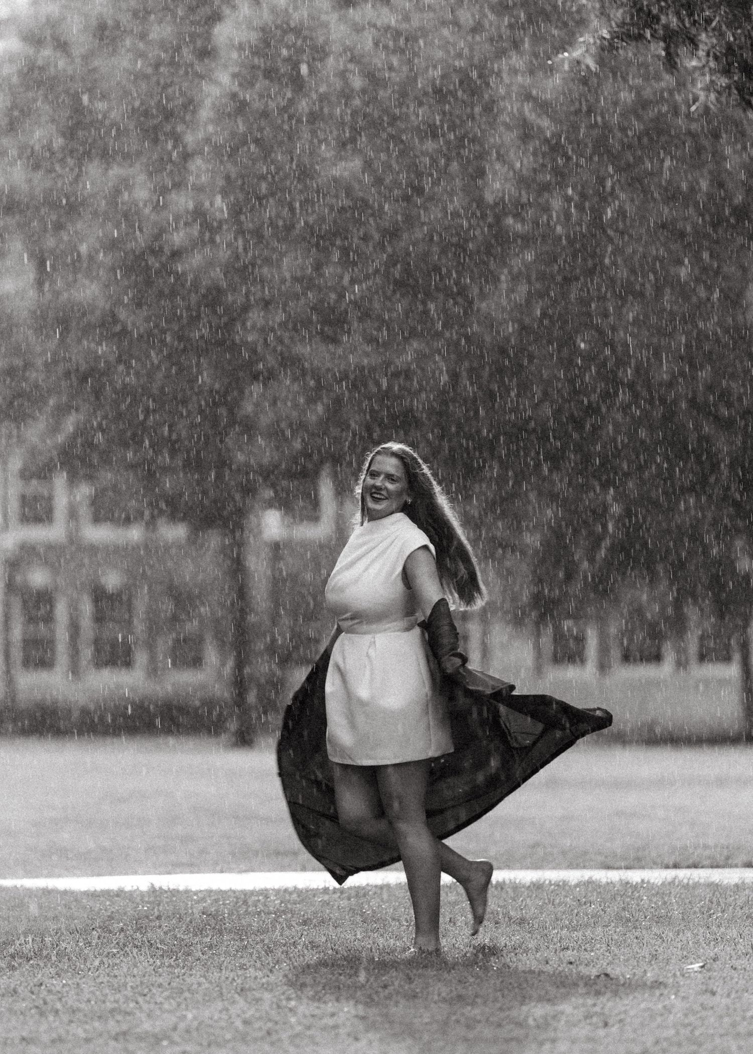 Smiling graduate twirls barefoot in rain with black gown flowing on campus lawn.