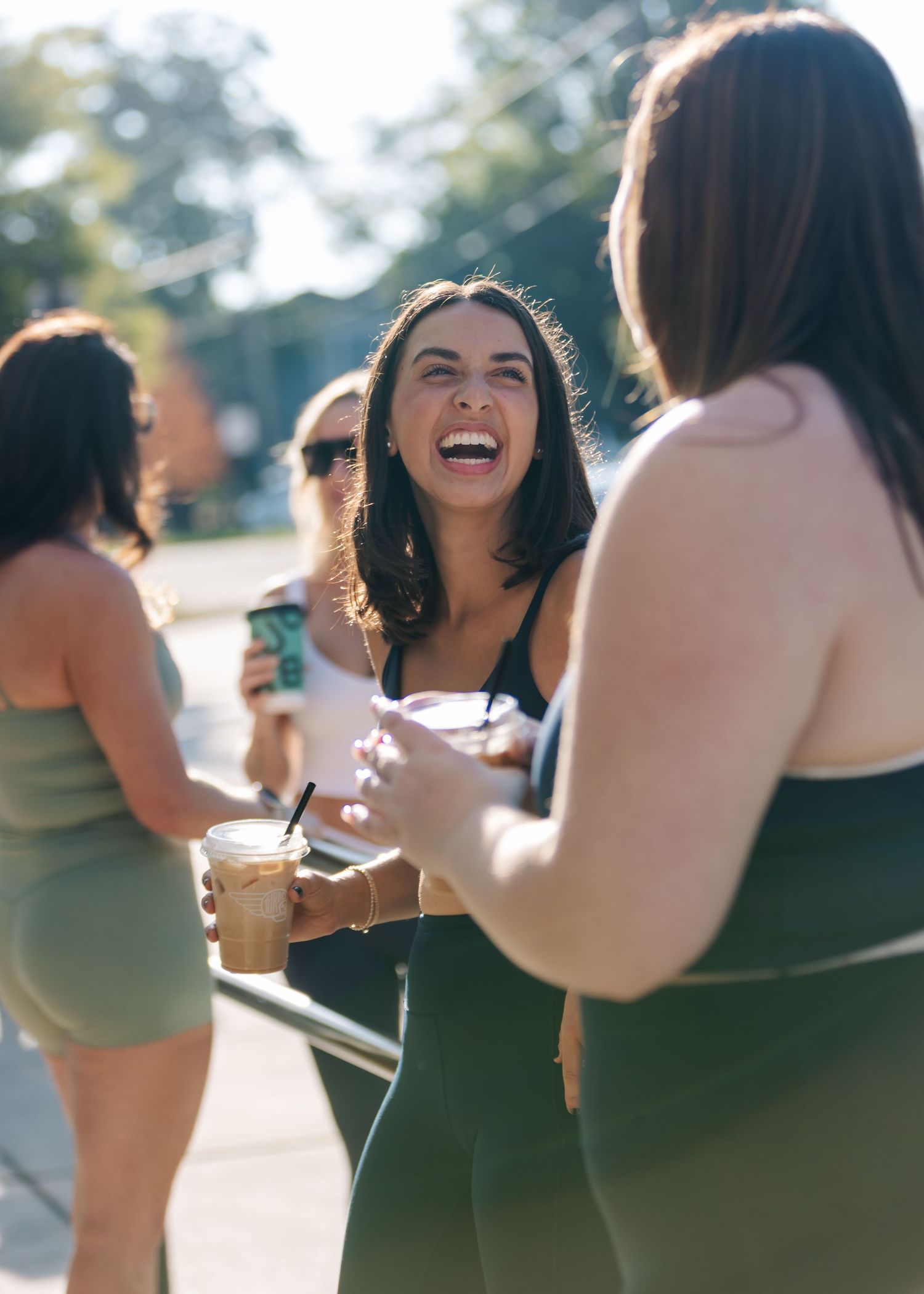 Women laughing and socializing outdoors holding iced coffee drinks in athletic wear on a sunny day.