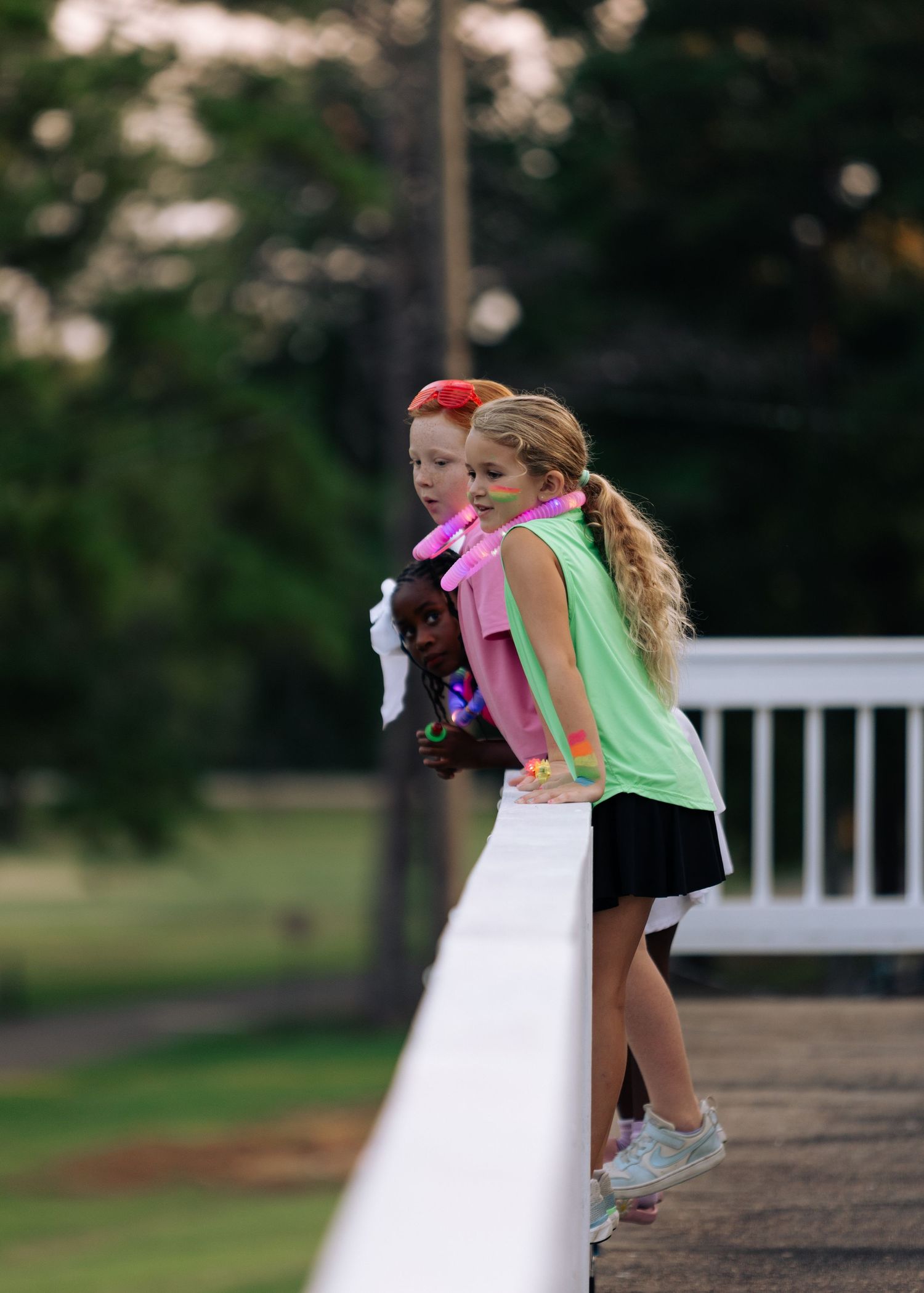 Two young girls lean against a white fence railing outdoors at dusk, one holding a stuffed animal.
