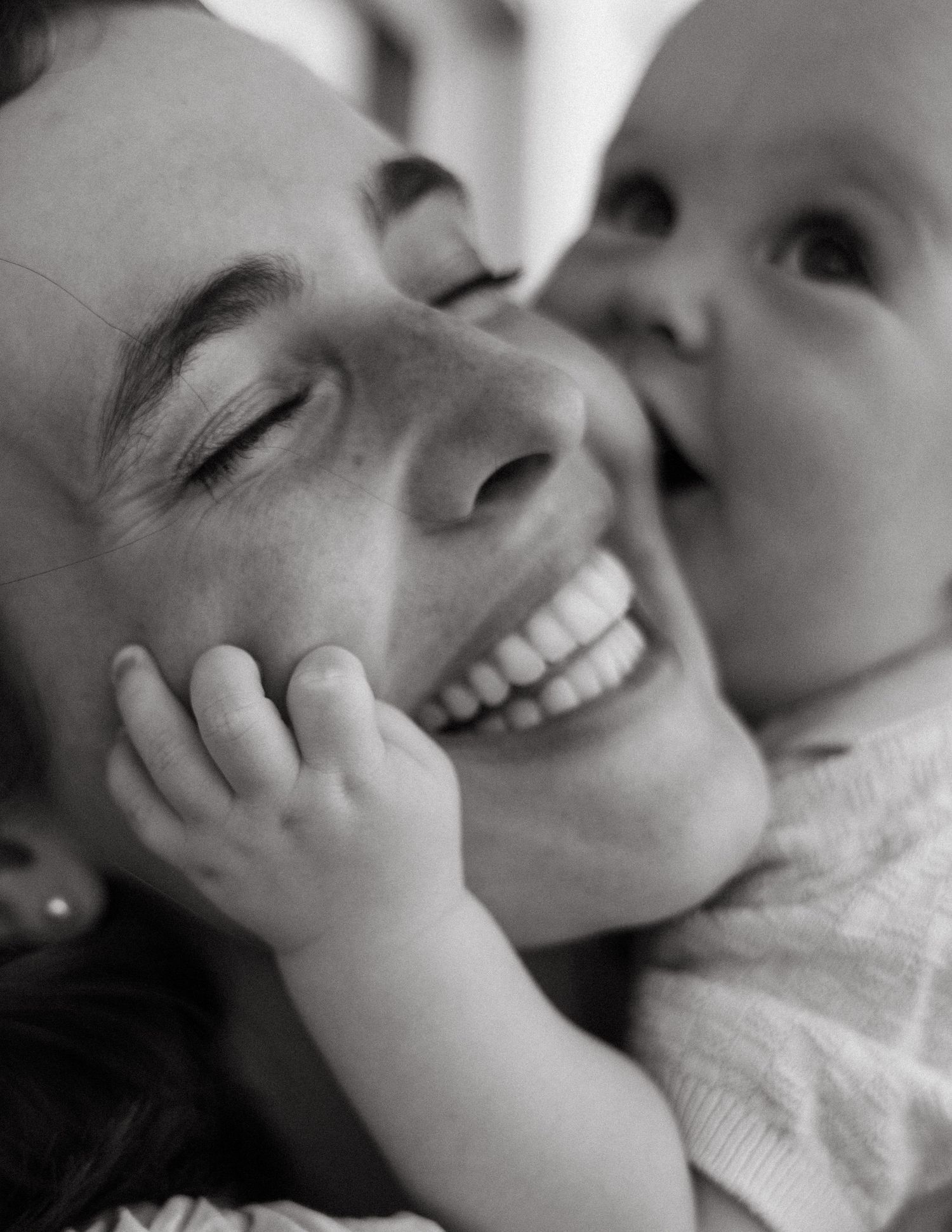 Black and white close-up of a smiling mother as her baby gently touches her face.