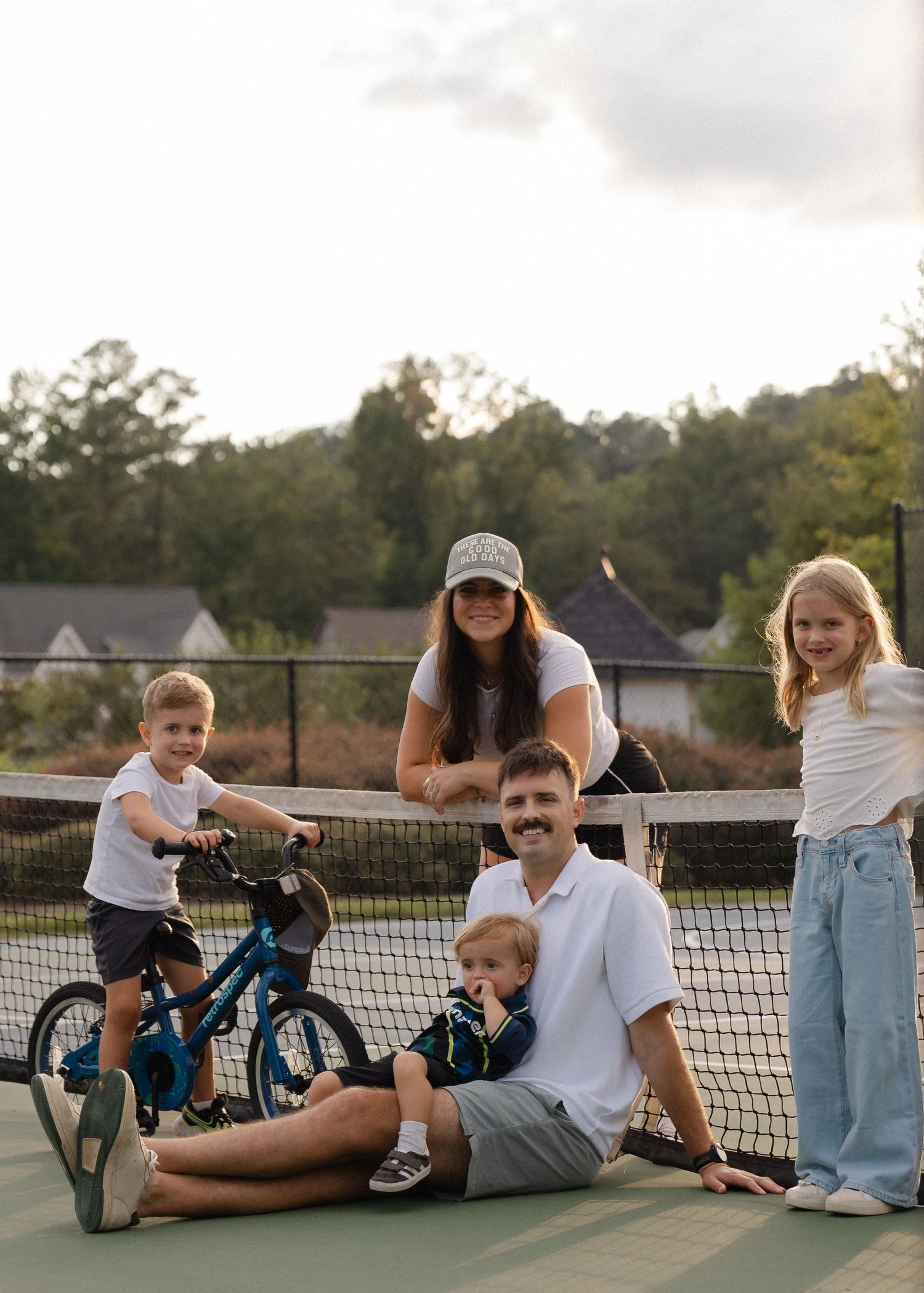 A family of five poses on a tennis court, with a boy on a bike, dad seated, and mom standing behind.