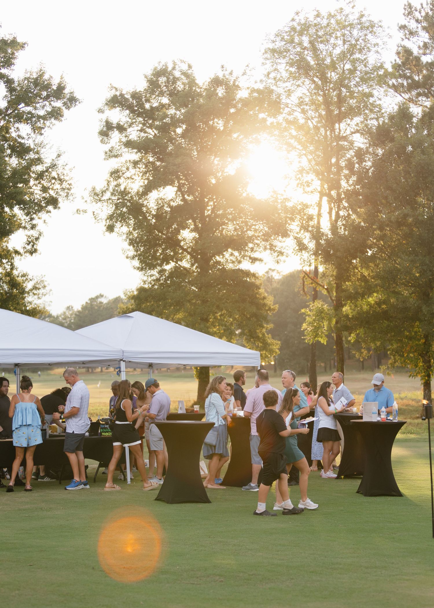 Guests mingle around black cocktail tables near white tents at an outdoor evening reception on a grassy lawn at golden hour.