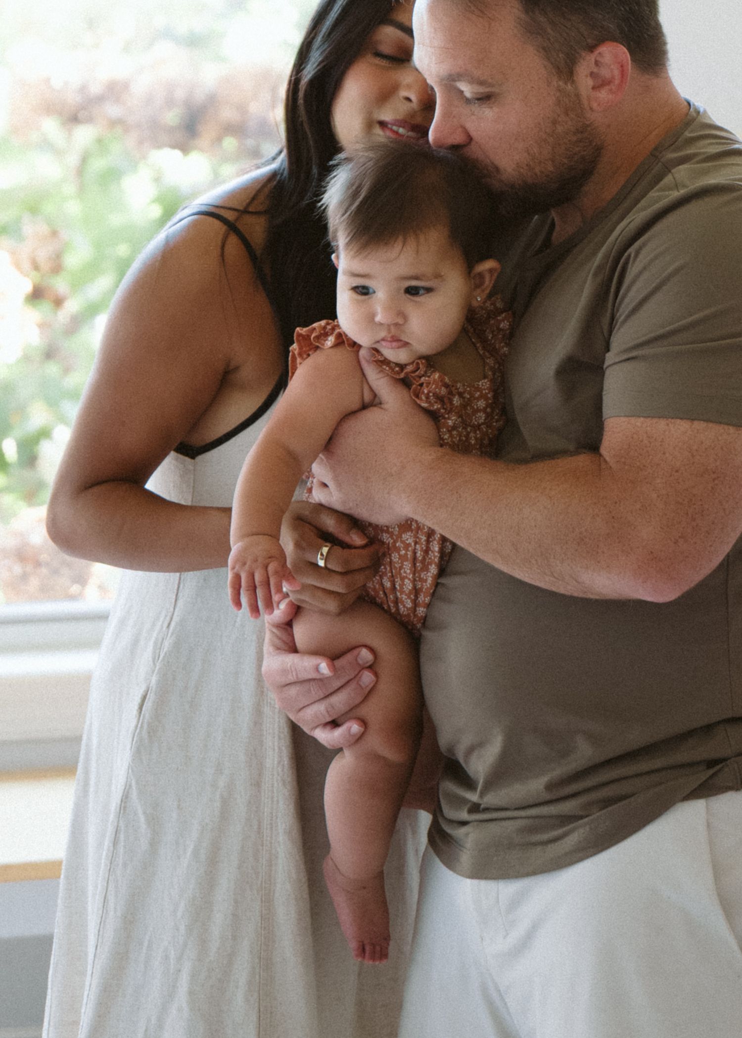 A couple tenderly holds their baby girl, sharing a loving family moment indoors near a bright window.