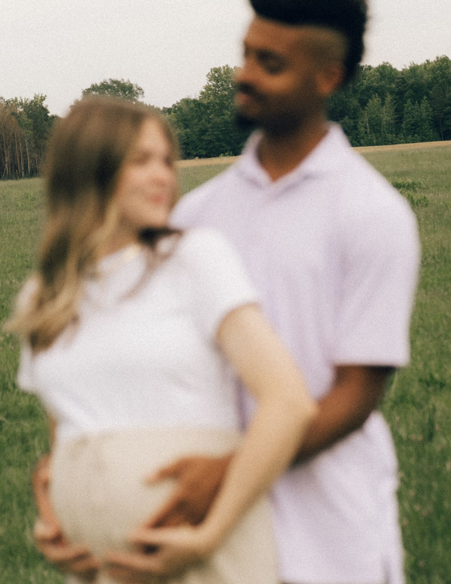 Soft focus maternity photo of couple embracing in a green field, man tenderly holds pregnant belly.