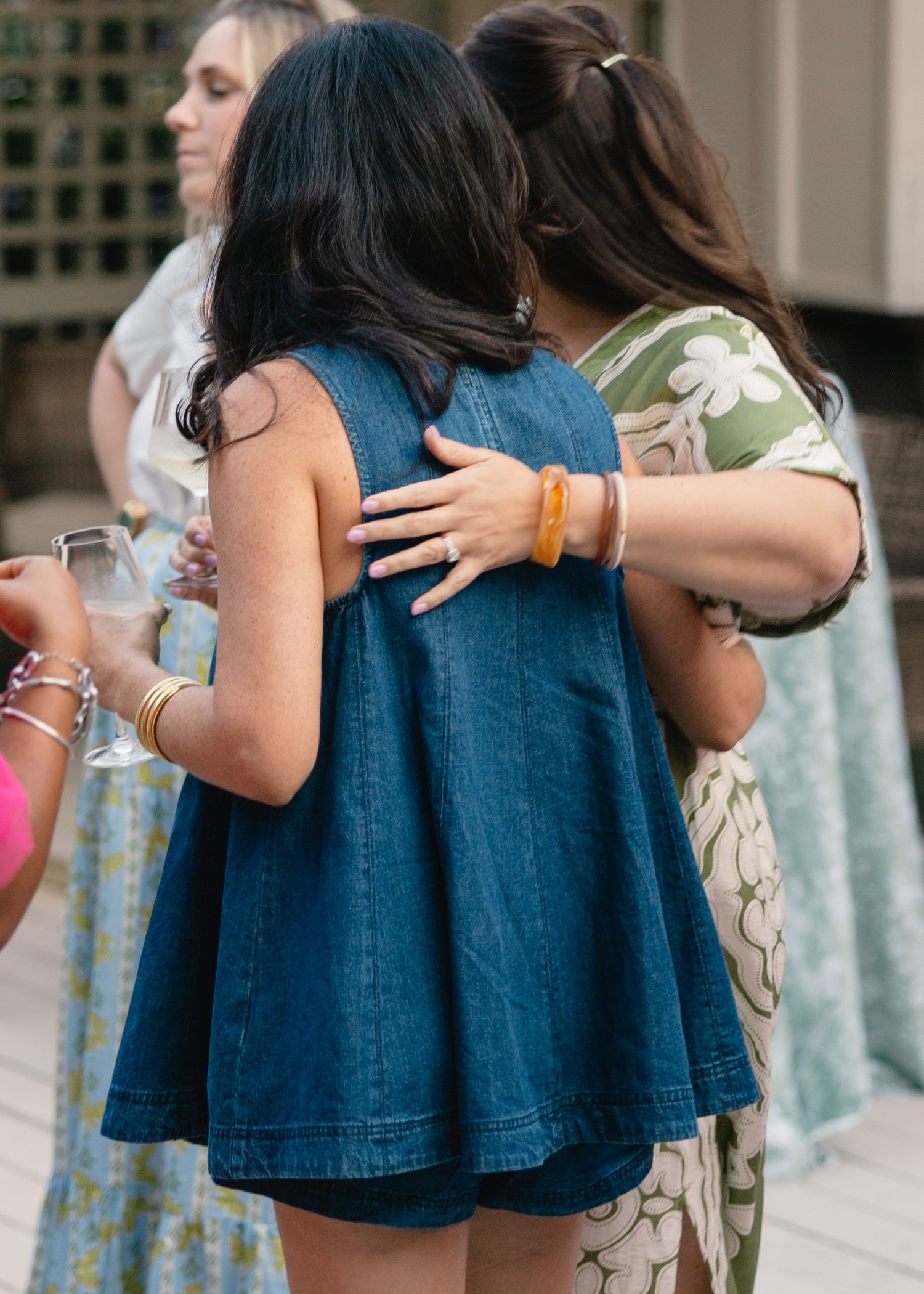 Women embracing at an outdoor summer party, one wearing a denim dress and another in a floral print top.