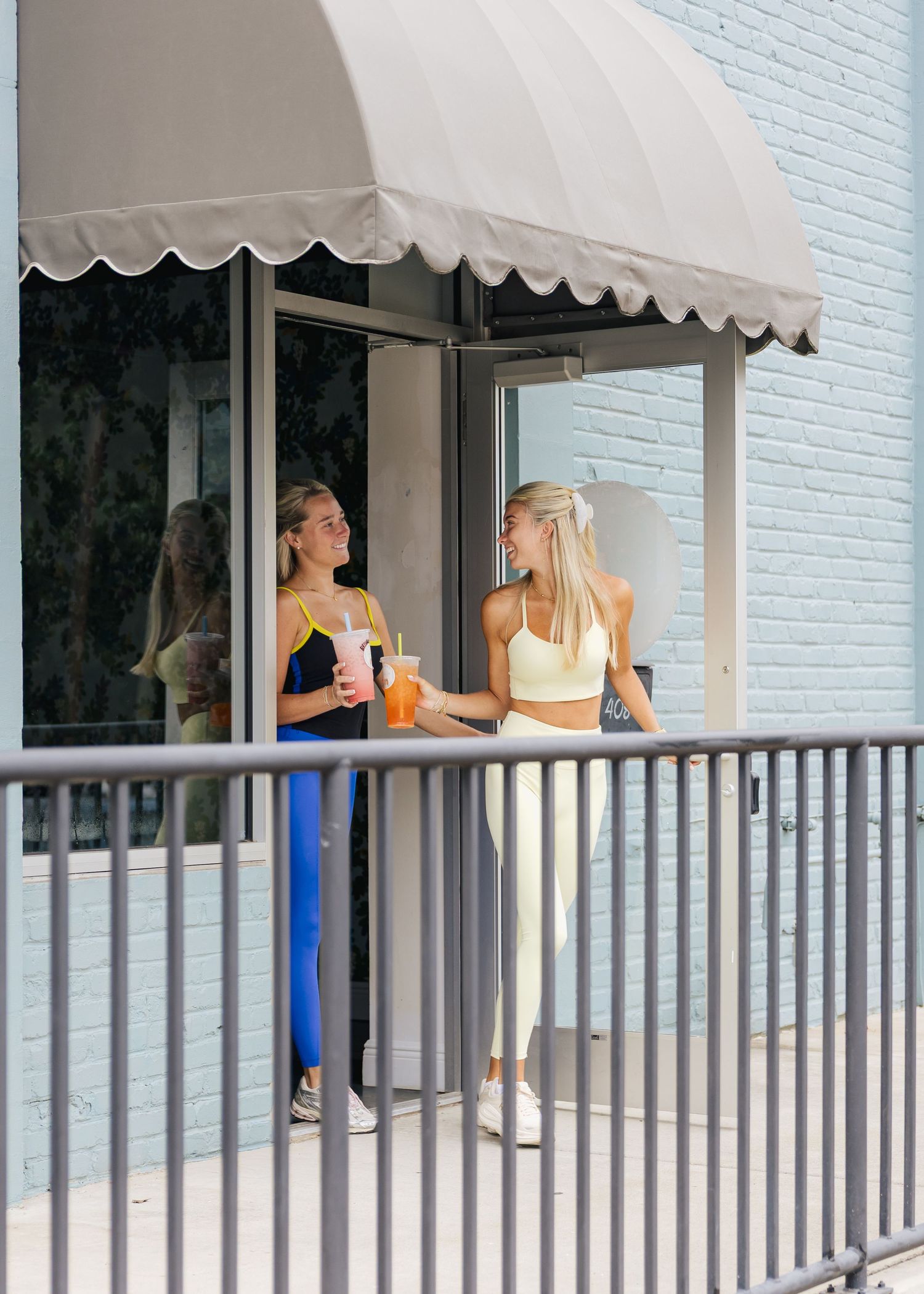 Two women in athletic wear holding colorful drinks outside a shop with a scalloped awning.