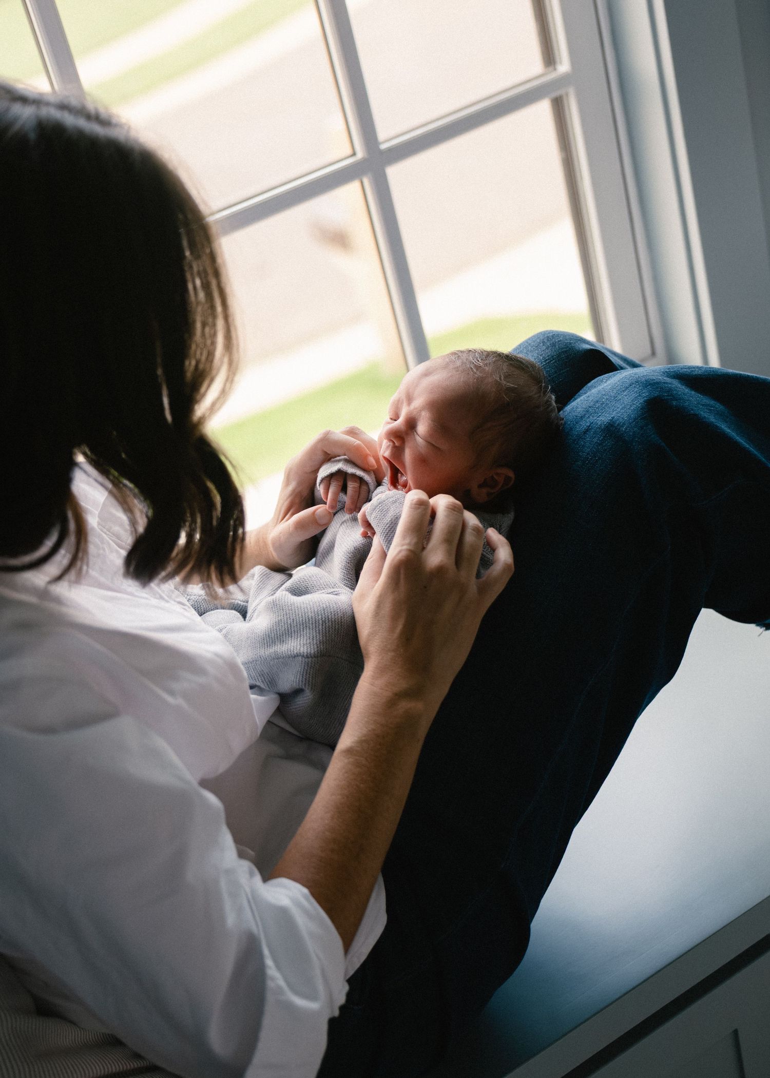 Mother in white shirt tenderly gazing at sleeping newborn baby by bright window.