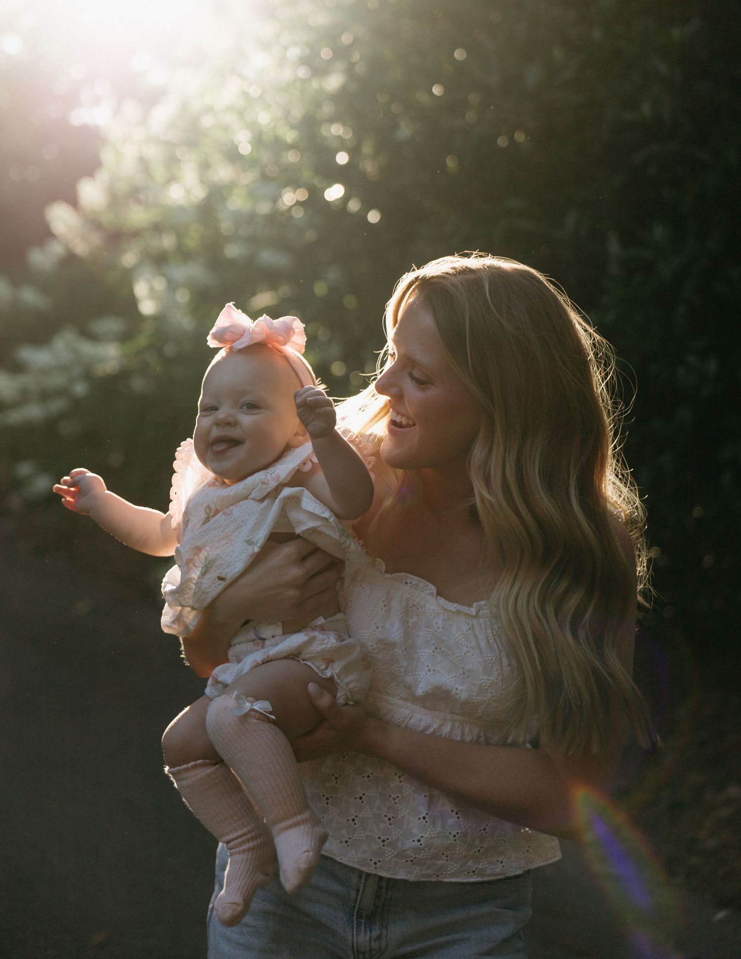 Young blonde mother holding smiling baby girl with pink bow in golden sunlight outdoors.