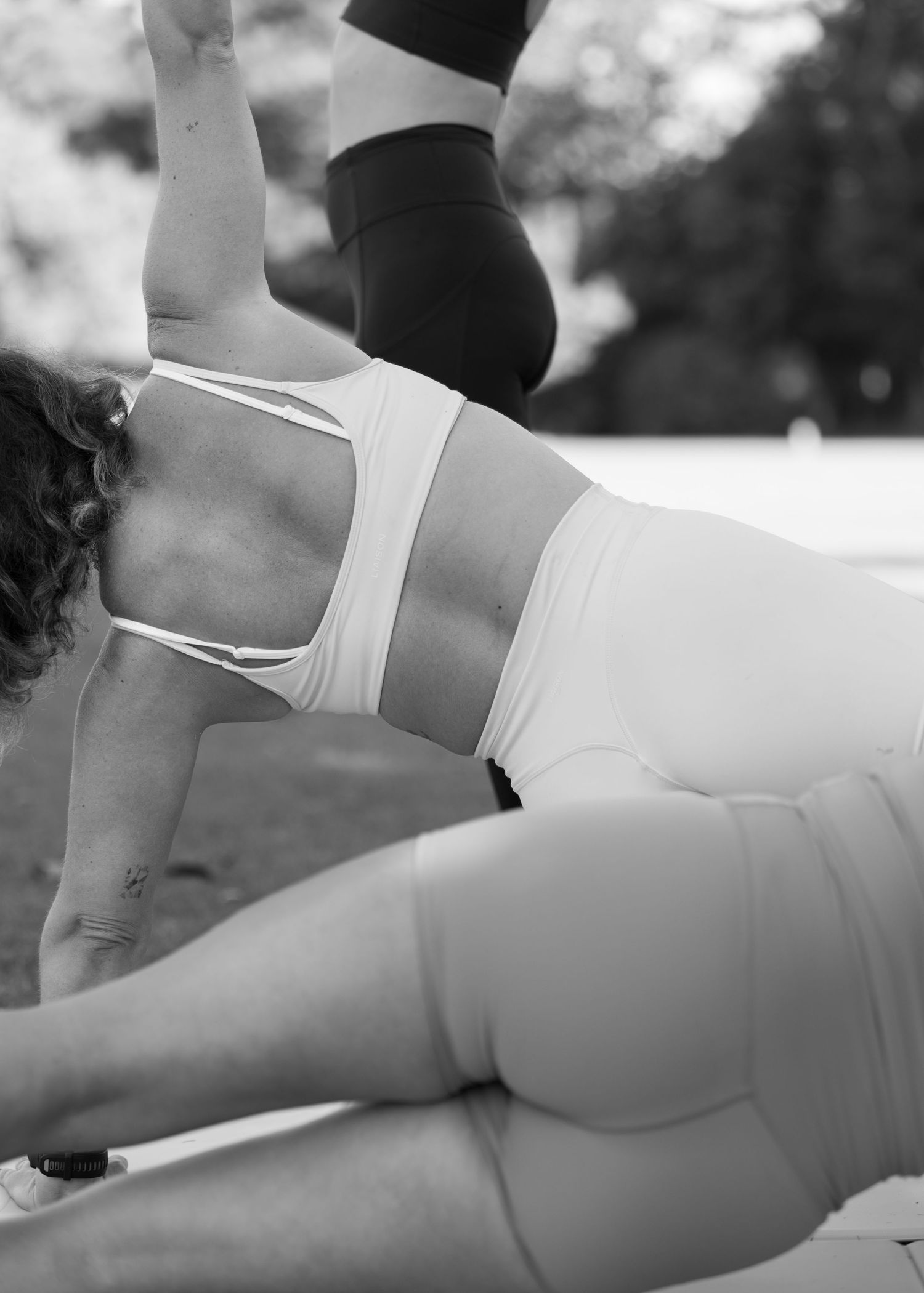 Black and white photo of two women exercising outdoors, one performing a side plank in athletic wear.