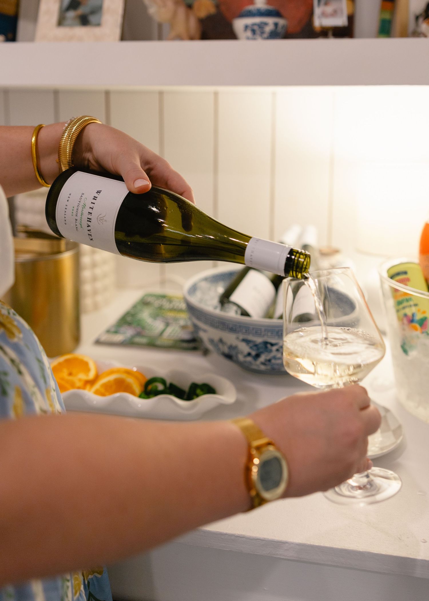 Woman pouring white wine into a stemmed glass at an elegant indoor party with fresh fruit and blue bowls.