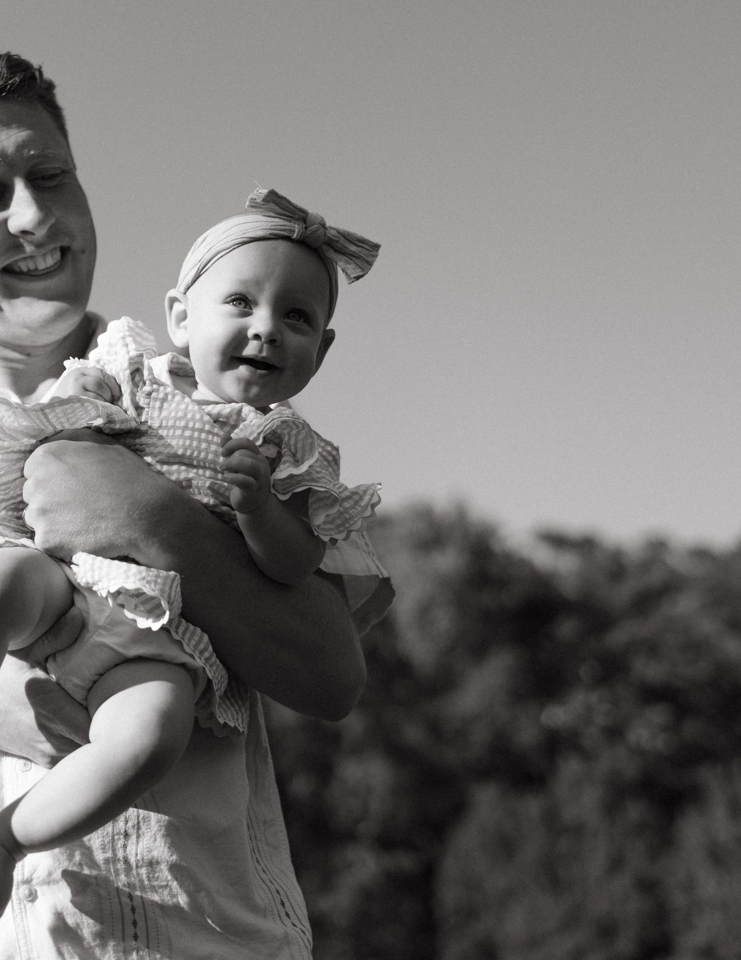 A smiling father holds his happy baby girl with a bow headband outdoors.