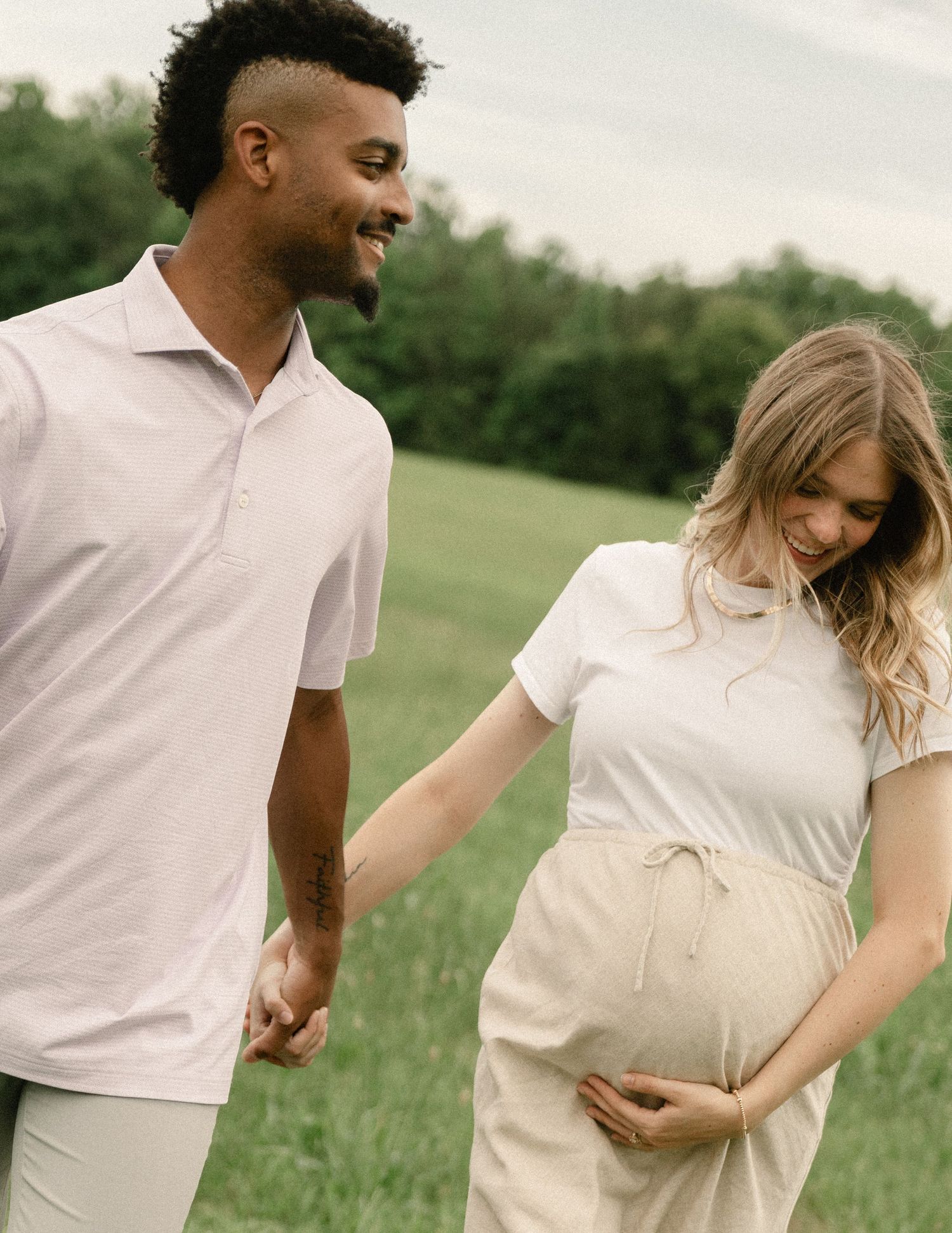 Smiling couple in a green field, man in polo shirt holds hands with pregnant woman in white maternity dress.