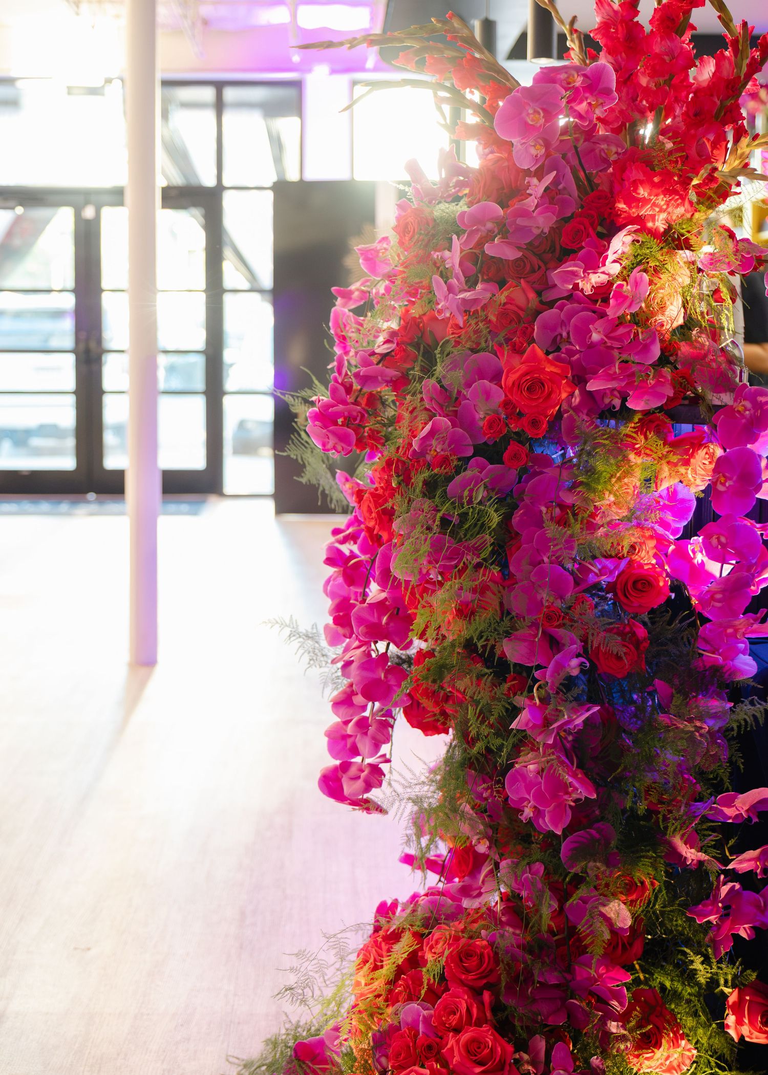 Lush floral installation of magenta orchids, red roses and green ferns arching over an event venue with glass doors and pink lighting.