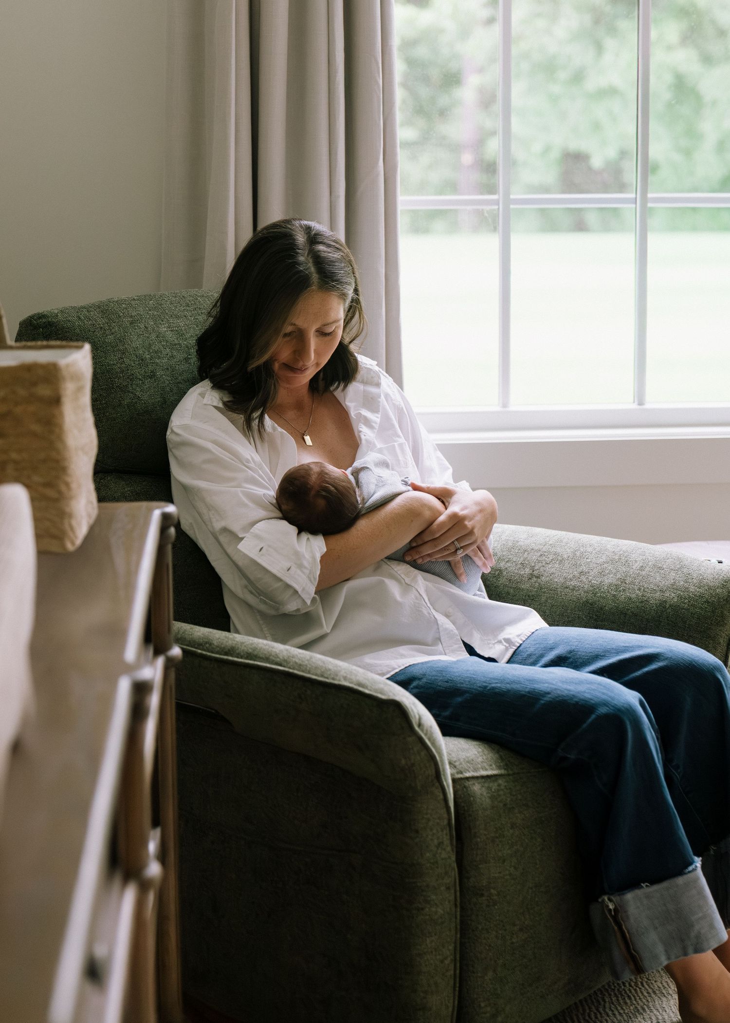 Mother in white shirt cradling newborn baby while seated in green armchair by window.