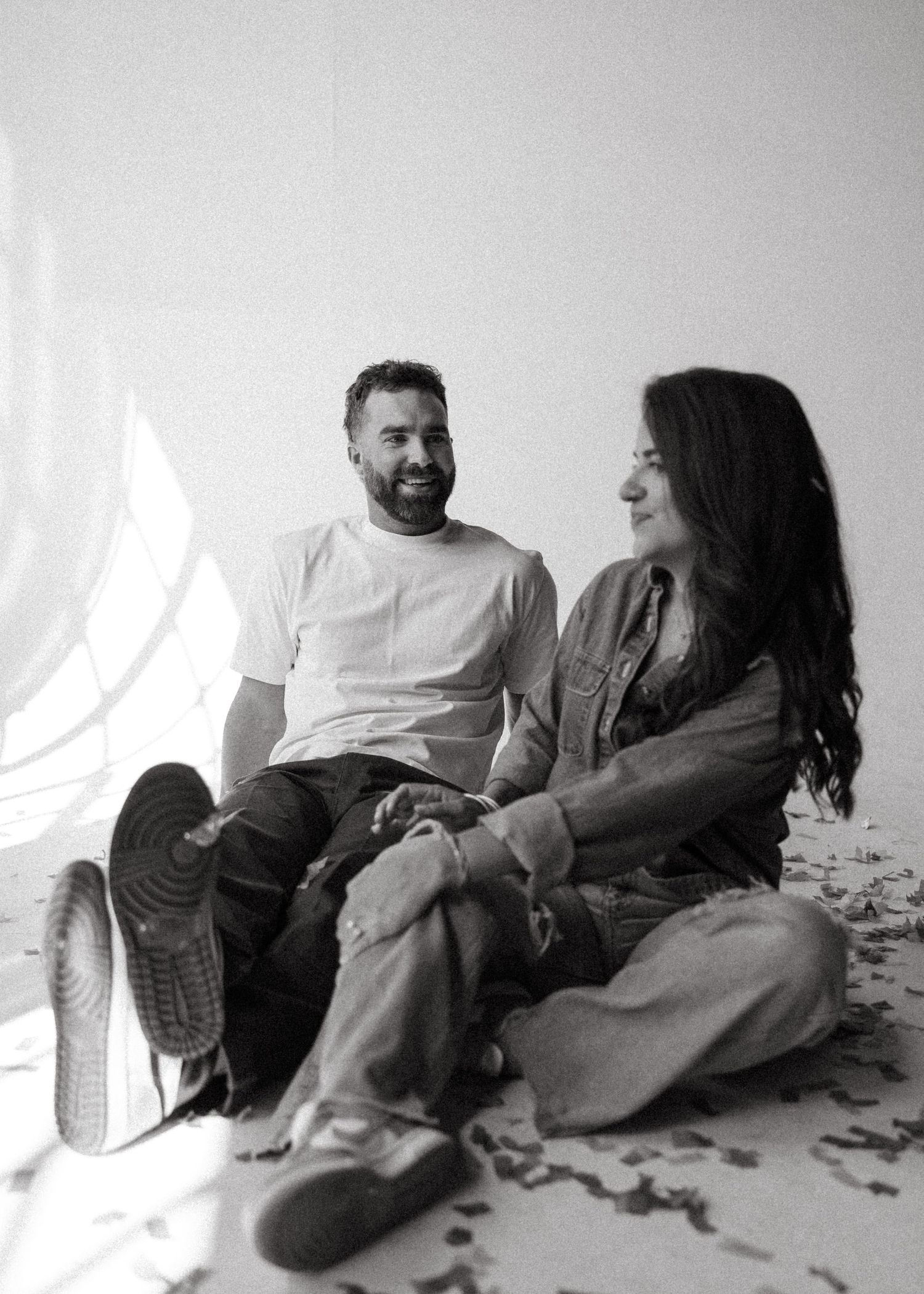 Black and white photo of a couple sitting on the floor laughing together in a bright studio setting.