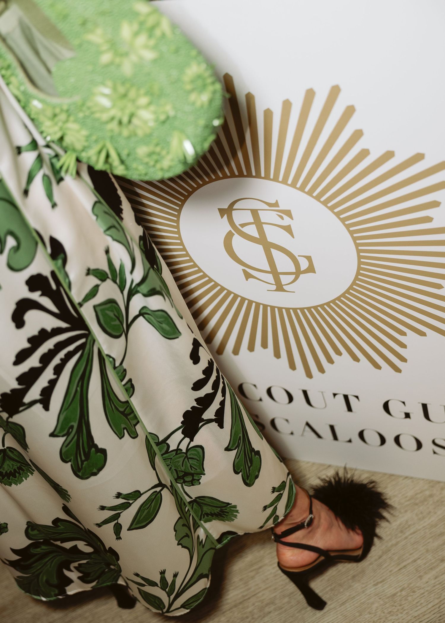 Woman in a green and cream leafy-print gown and black feathered heels stands beside a gold sunburst monogram sign at an event.