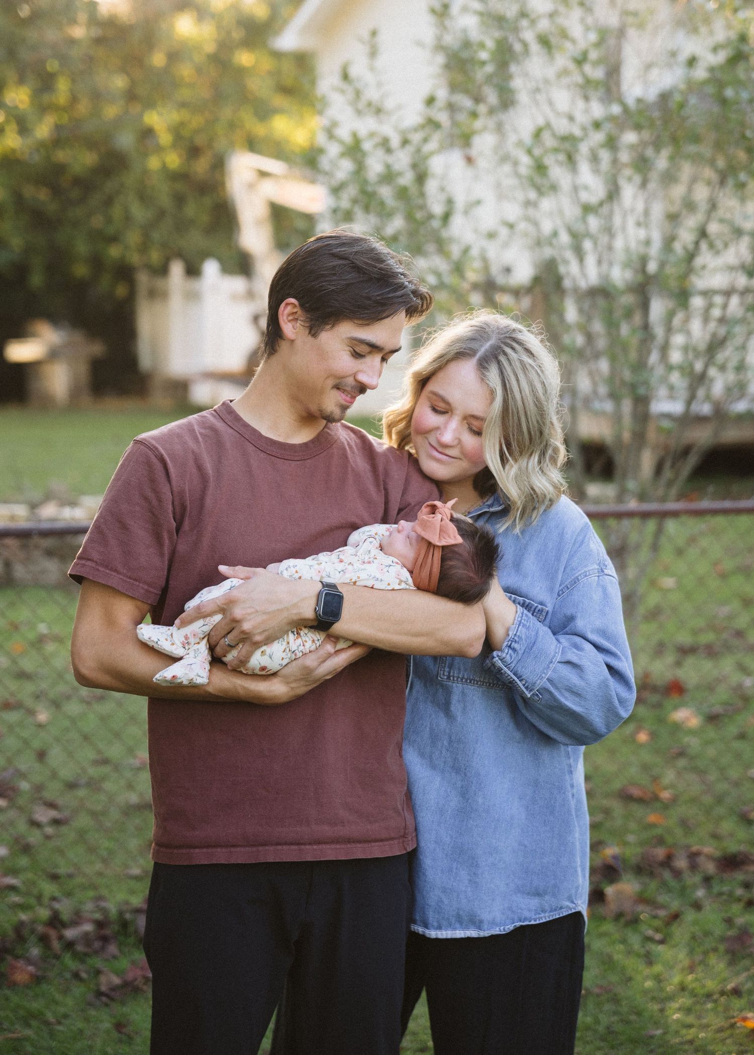A smiling couple stands in a leafy backyard cradling a sleeping newborn baby in a floral outfit and orange bow headband.