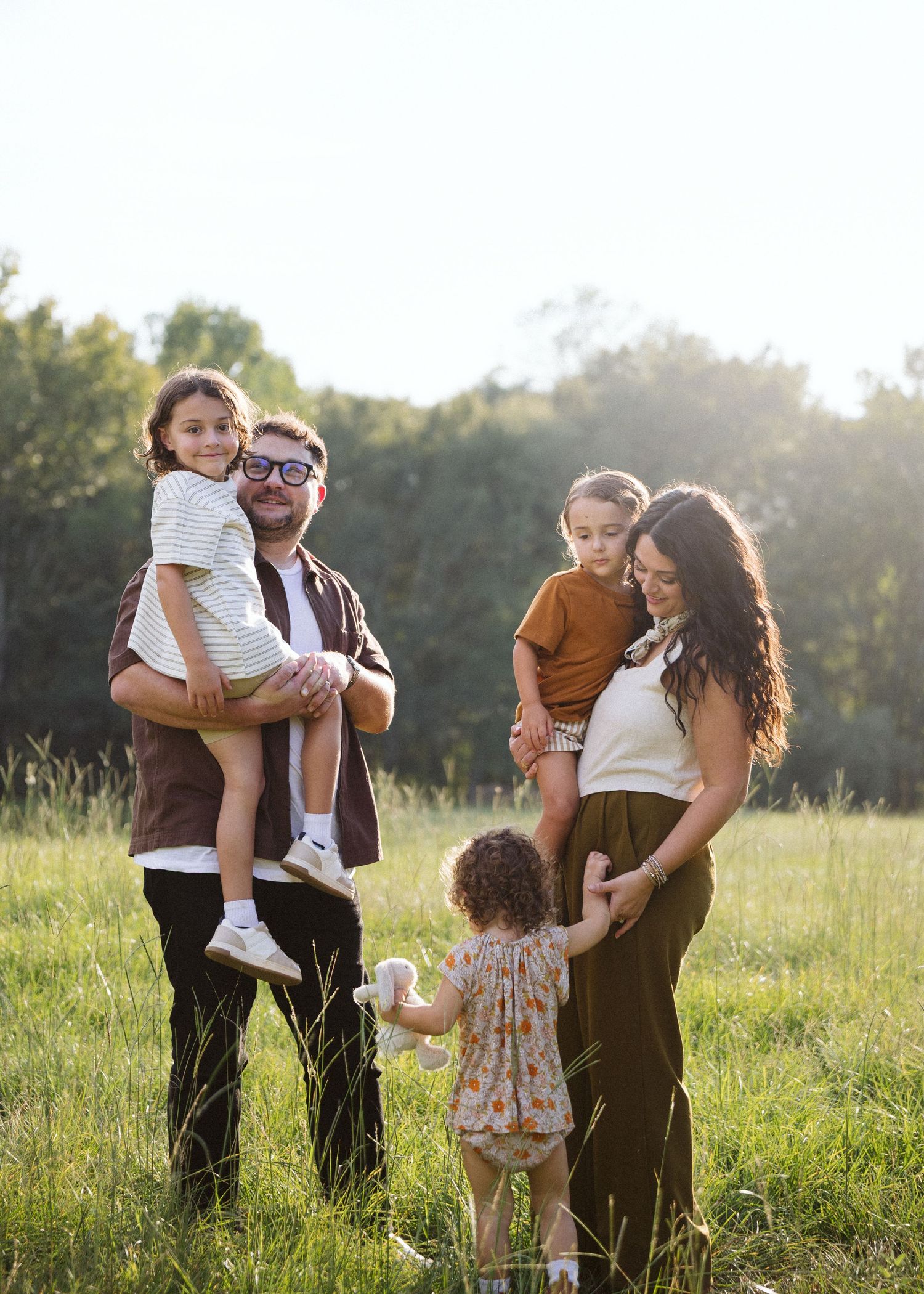 Family of five in earth-tone outfits standing in a sunlit grassy field, parents holding two children while a toddler holds a stuffed toy.